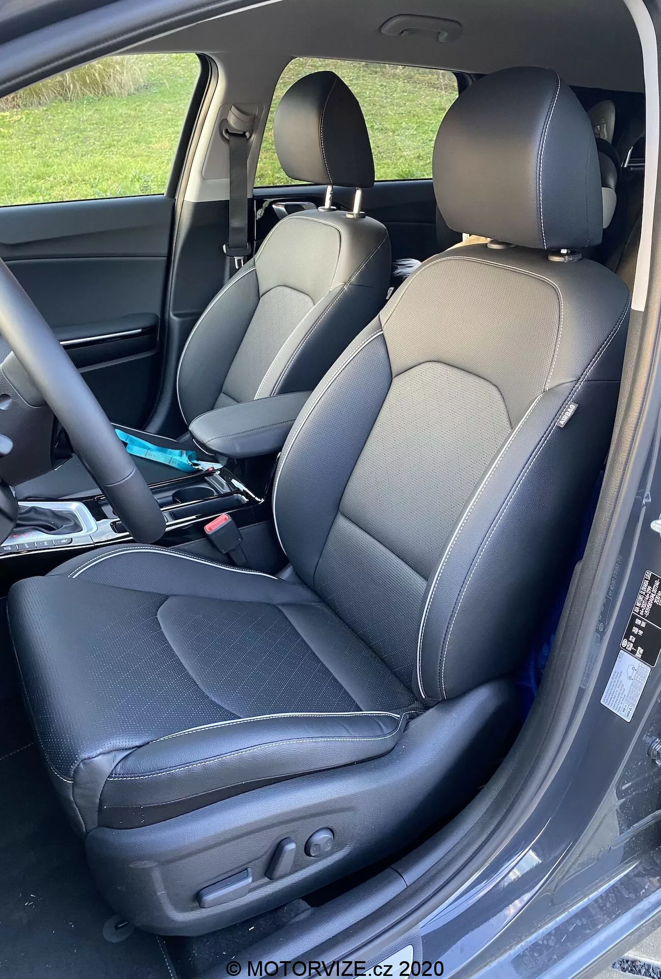 Imagem aproximada do interior do Kia Ceed 2018, mostrando os assentos do motorista e do passageiro com estofamento em couro de duas tonalidades e costuras contrastantes, design esportivo com apoios laterais, e ajustes elétricos no assento do motorista para controle do ângulo do encosto e da posição do assento. Vincos leve indicam uso e diversos itens são visíveis no assento do passageiro.