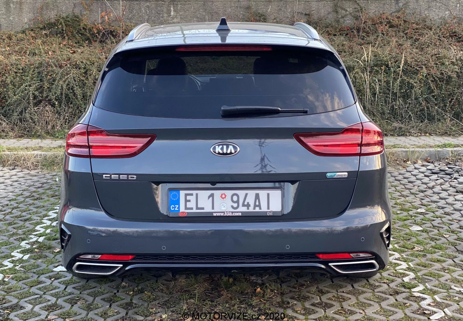 Vista traseira de um Kia Ceed 2018 mostrando toda a secção traseira com emblemas de marca e modelo, modernas luzes traseiras LED, janela traseira, saídas de escape duplas, para-choques traseiro com refletores, spoiler de teto, antena e sensores de estacionamento traseiros. A imagem é clara, tirada ao nível dos olhos com iluminação natural em condições de céu nublado.