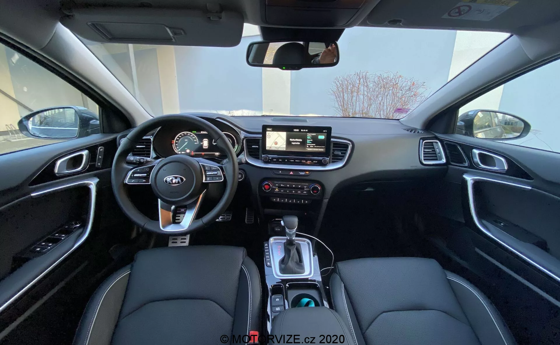 Uma visão ampla do interior do Kia Ceed 2018 a partir do banco traseiro, mostrando a área do motorista, incluindo o volante com controles montados, painel de instrumentos com medidores digitais e analógicos, sistema de infoentretenimento central com tela sensível ao toque, controles manuais para infoentretenimento e clima, e console central com alavanca de câmbio, seletor de modo de condução, porta-copos e um interior preto com assentos de couro sintético com costura branca e acabamento de alumínio escovado.
