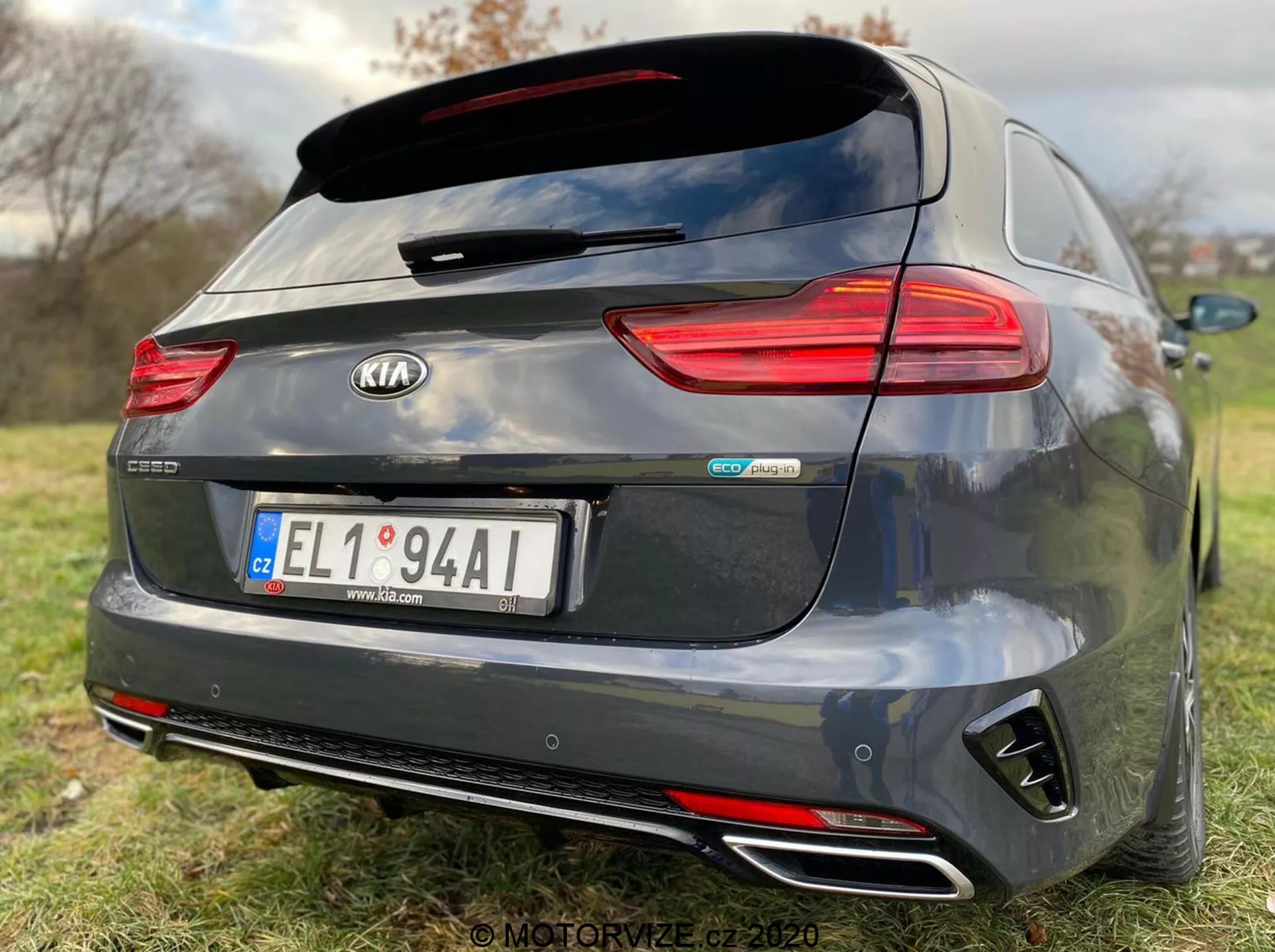 Vista traseira de um modelo Kia Ceed Proceed 2018 cinza escuro, mostrando características detalhadas como luzes traseiras horizontais de LED, um spoiler traseiro montado no teto, pontas de escapamento duplas cromadas, para-choques traseiro em estilo difusor preto e emblemas centrais incluindo 'Kia' e 'GT Line'. A câmera de ré acima da placa central sugere assistência ao estacionamento, enquanto a pouca profundidade de campo enfatiza a marca e os identificadores do modelo do carro em um fundo gramado.