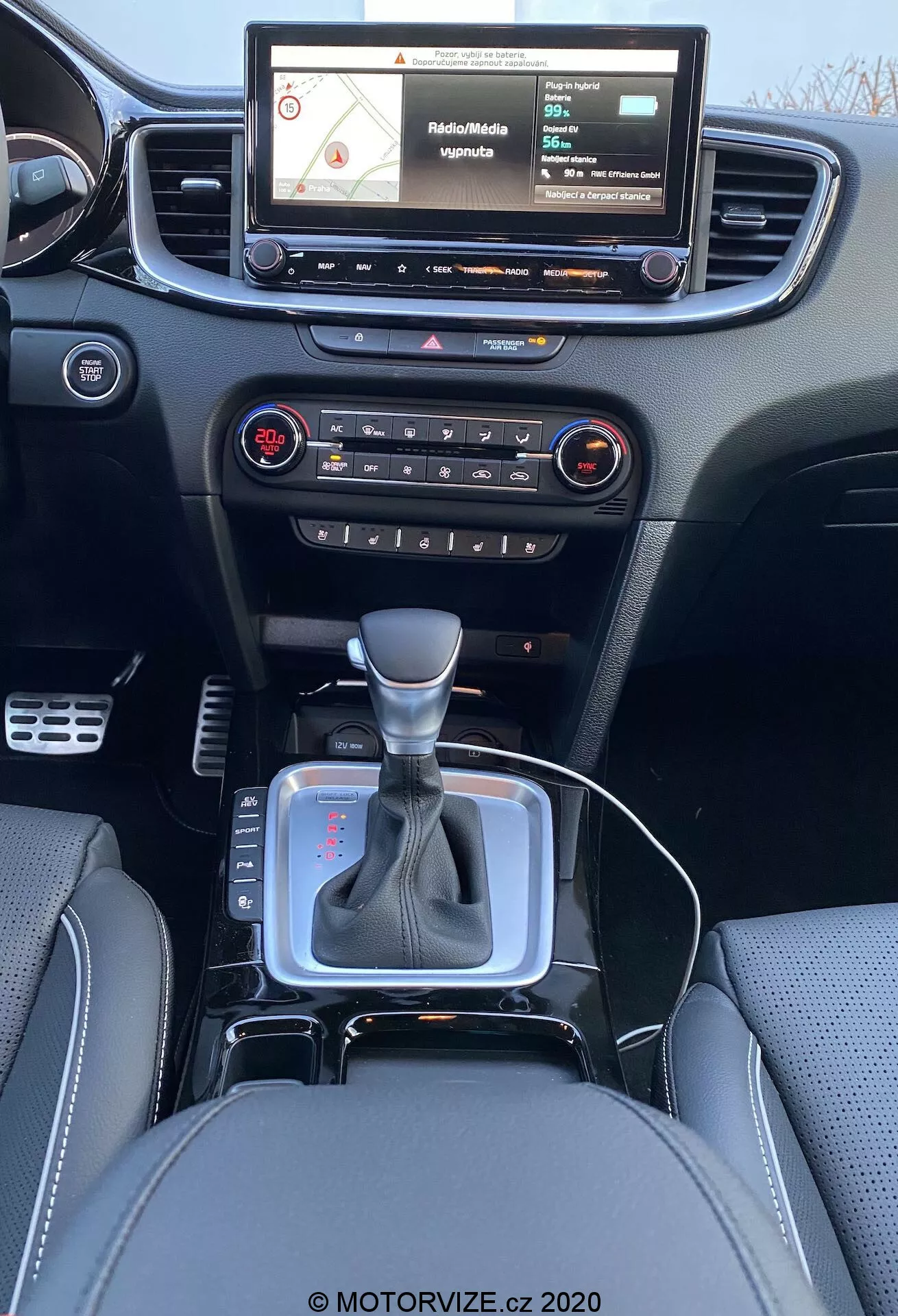 Vista interior de um Kia Ceed 2018 a partir da perspectiva do banco traseiro, mostrando um painel moderno com um sistema de infotainment com tela sensível ao toque exibindo um mapa de navegação, botões físicos e botões para controle de áudio e clima, ventilador central, alavanca de câmbio em posição neutra com detalhe prateado e capa de couro preta, pequeno espaço de armazenamento próximo à alavanca de câmbio possivelmente para tomada de 12V ou portas USB, bancos de couro com costuras detalhadas.