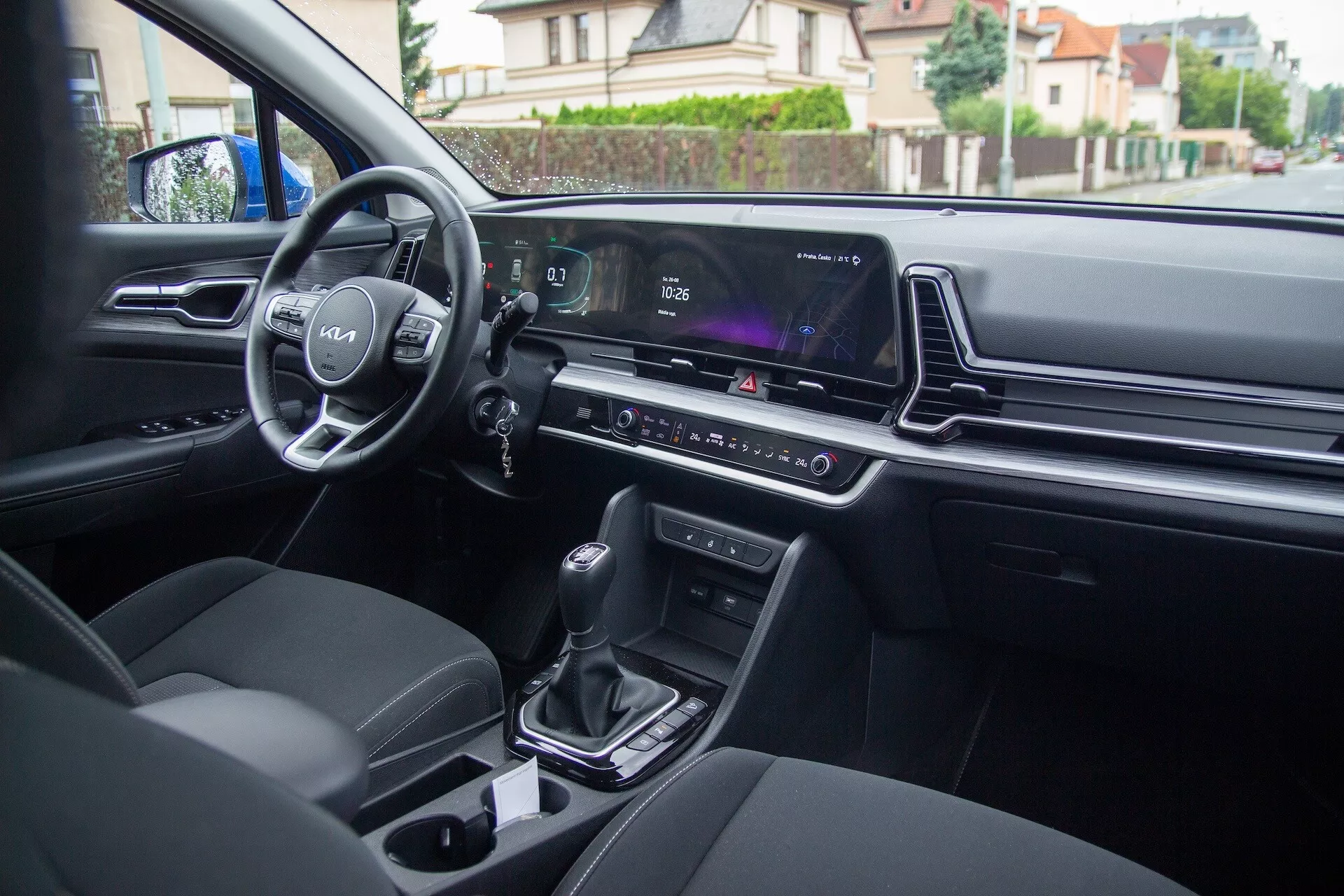 Vista interior do Kia Sportage 2022 a partir do banco traseiro mostrando os controles na porta do motorista, volante com logotipo e controles montados, painel de instrumentos digital, tela de infotenimento, controle climático, alavanca de mudanças automática, porta-copos e assentos modernos estofados