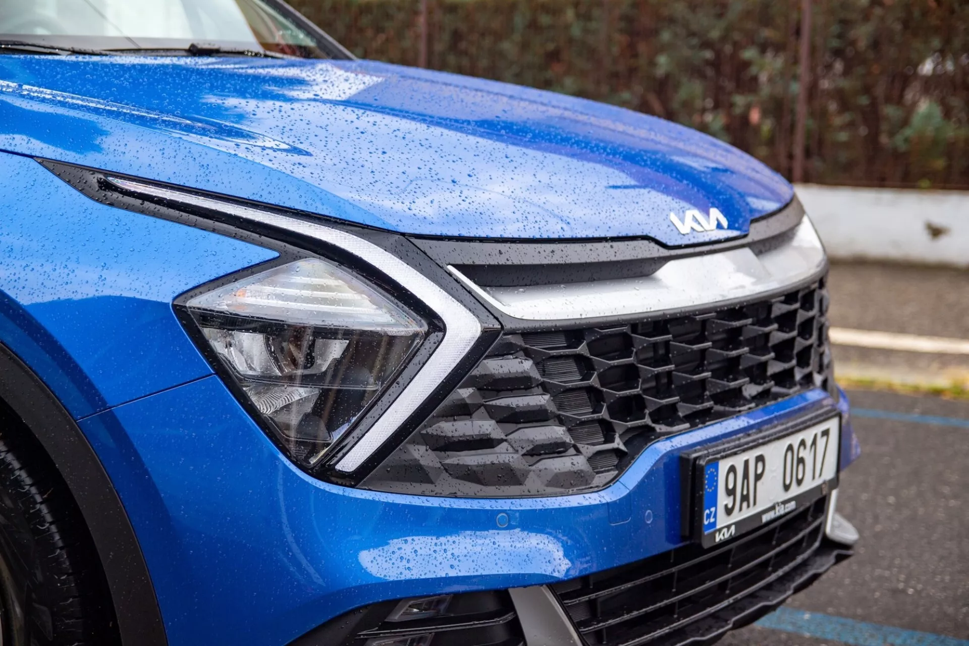 Vista detalhada de um Kia Sportage azul 2022 focada no quarto dianteiro esquerdo em condições úmidas, destacando o design dinâmico do capô, faróis LED, grade de favo de mel e tecnologia de sensores avançada