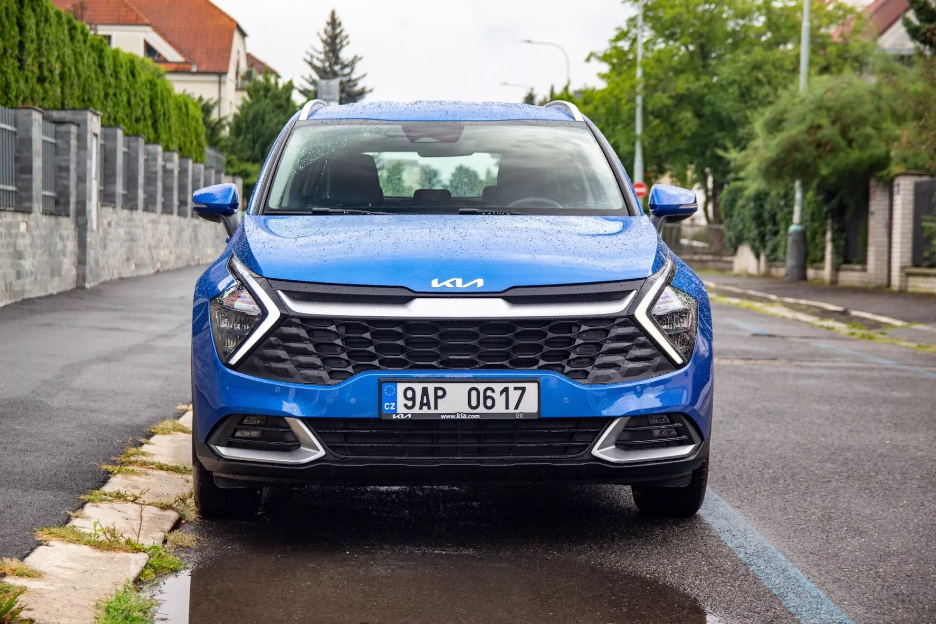 Vista frontal de um Kia Sportage azul de 2022, estacionado em uma estrada suburbana, exibindo sua grade moderna, faróis elegantes, emblema Kia em cromo e design dianteiro dinâmico