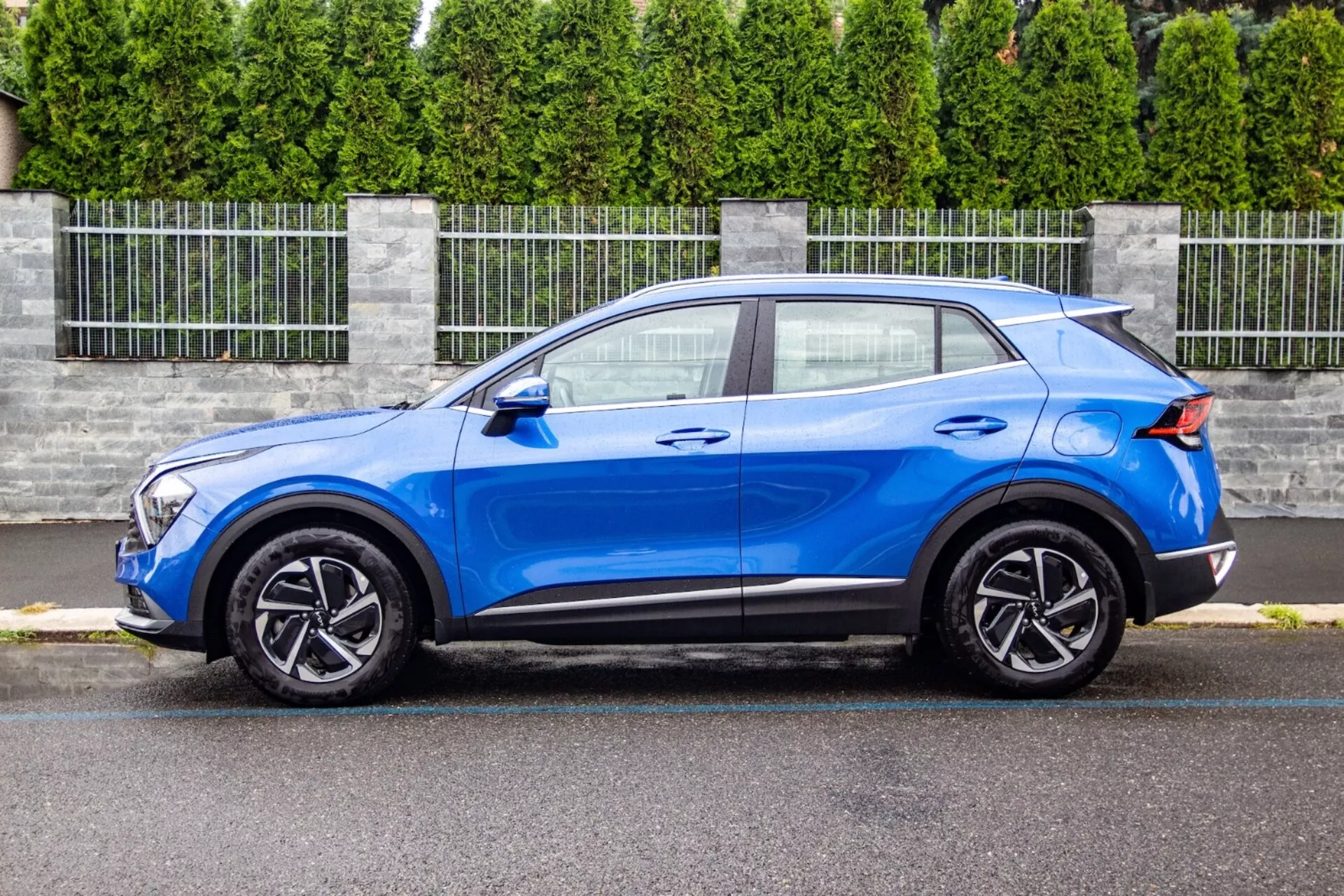 SUV crossover Kia Sportage 2022 em azul brilhante, exibindo seu design elegante e compacto com uma linha de teto inclinada ao estilo de um coupé, capô contornado, unidades de farol LED, rodas de liga de cinco raios e melhorias aerodinâmicas como um spoiler traseiro, em um ambiente suburbano.
