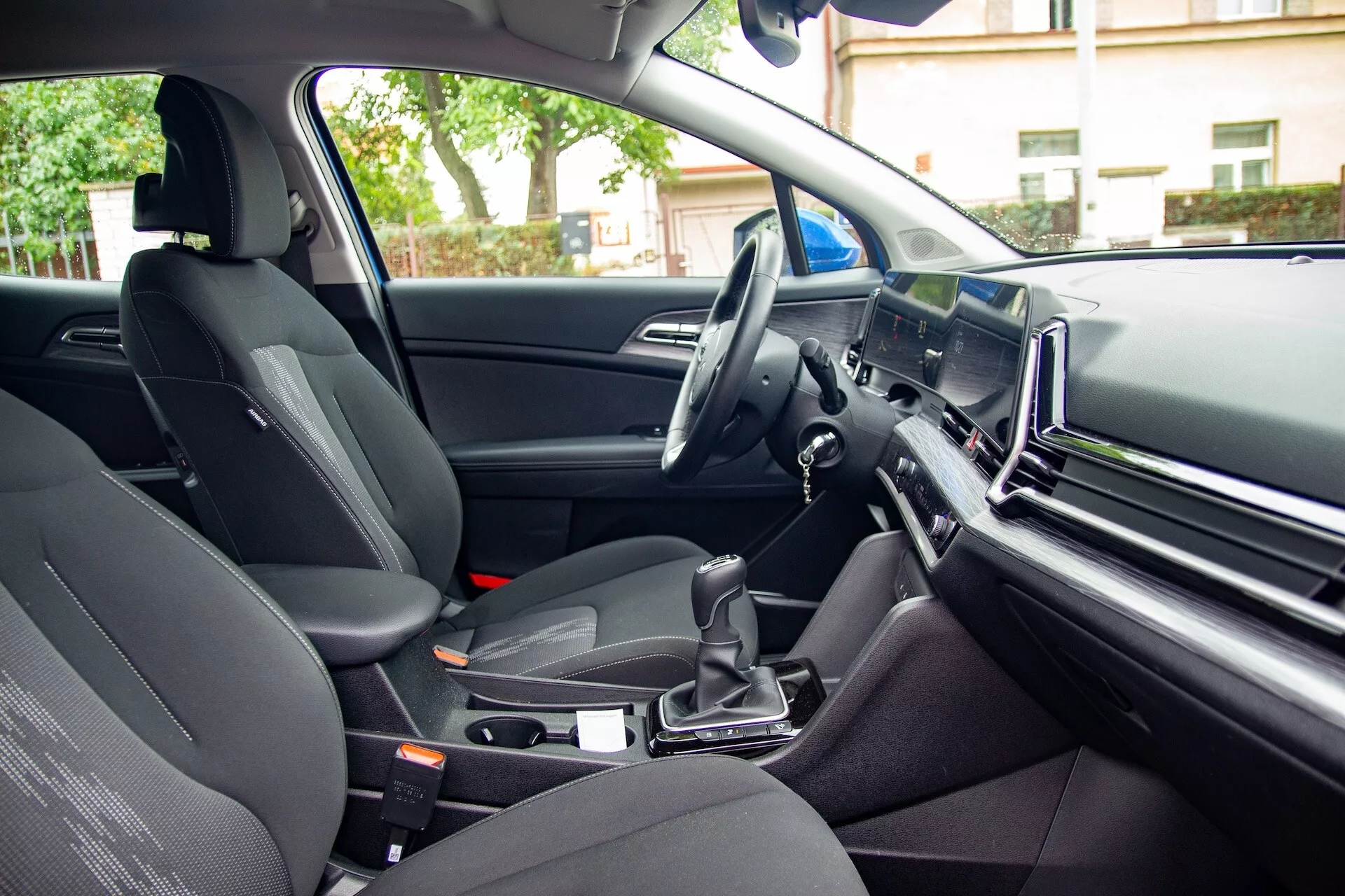 Vista interior do Kia Sportage 2022 a partir da perspectiva do passageiro traseiro mostrando o assento do passageiro dianteiro, console central com alavanca de câmbio automático, volante à esquerda e uma grande tela de infotenimento no painel