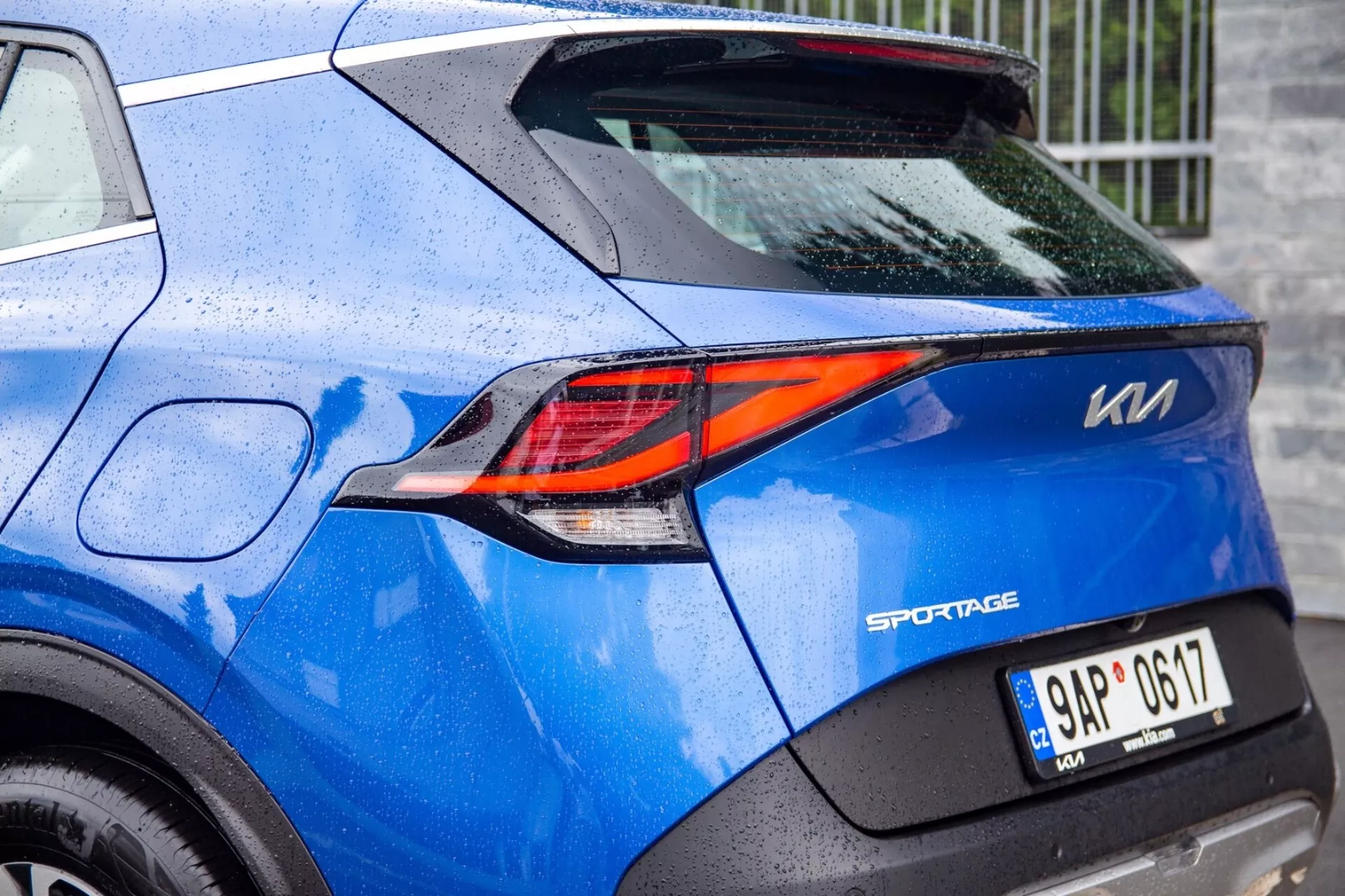 Vista parcial de um Kia Sportage azul 2022 destacando a sofisticada lanterna traseira LED em forma de Z e detalhes de design traseiro, incluindo o emblema do modelo, placa de licença e pintura salpicada de chuva