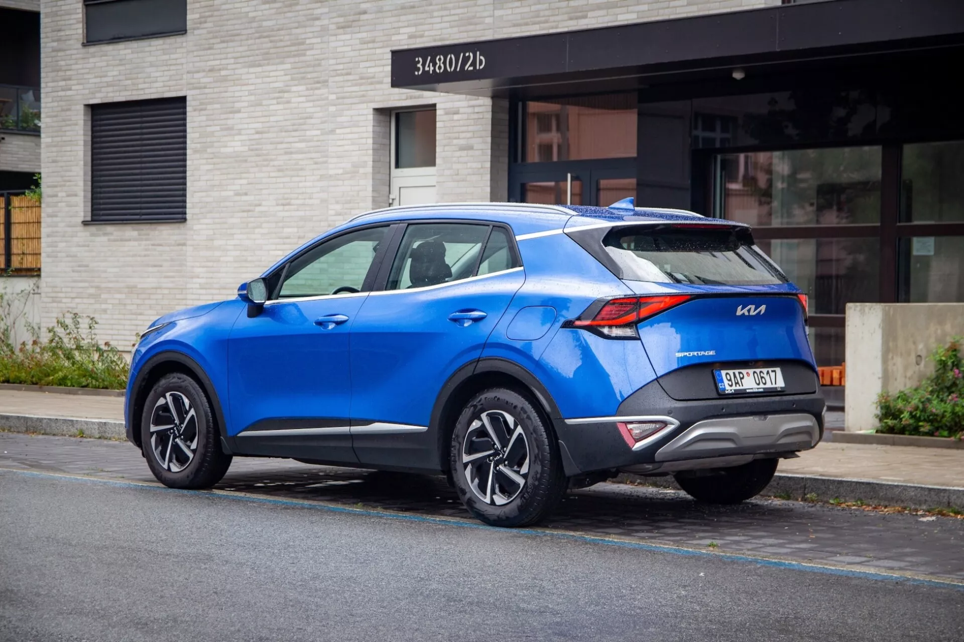 SUV crossover moderno Kia Sportage 2022 estacionado em uma rua da cidade, mostrando vista traseira de três quartos com luzes traseiras distintas, spoiler esportivo no teto, rodas de liga leve multirradiadas, cor azul vibrante e possíveis características tecnológicas modernas.