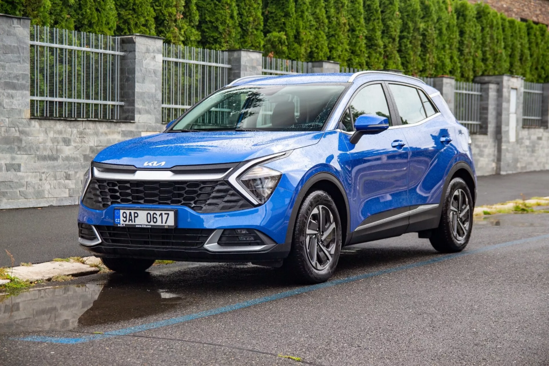 SUV compacto Kia Sportage 2022 em azul estacionado em uma rua de asfalto molhado mostrando grade frontal, faróis, para-choque, capô e perfil do lado do motorista a partir de um ângulo frontal de três quartos com perspectiva baixa para destacar o design do veículo e características tecnológicas