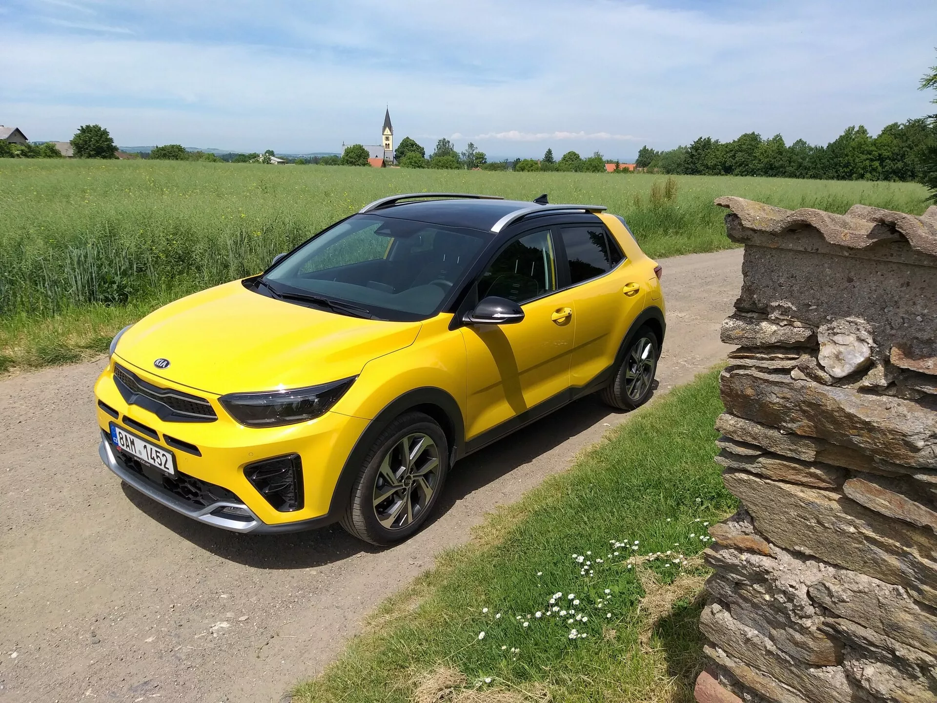 SUV compacto Kia Stonic 2017 amarelo brilhante com teto preto, exibindo seu design elegante e contornos dinâmicos a partir de um ângulo frontal de três quartos em uma estrada de terra rural