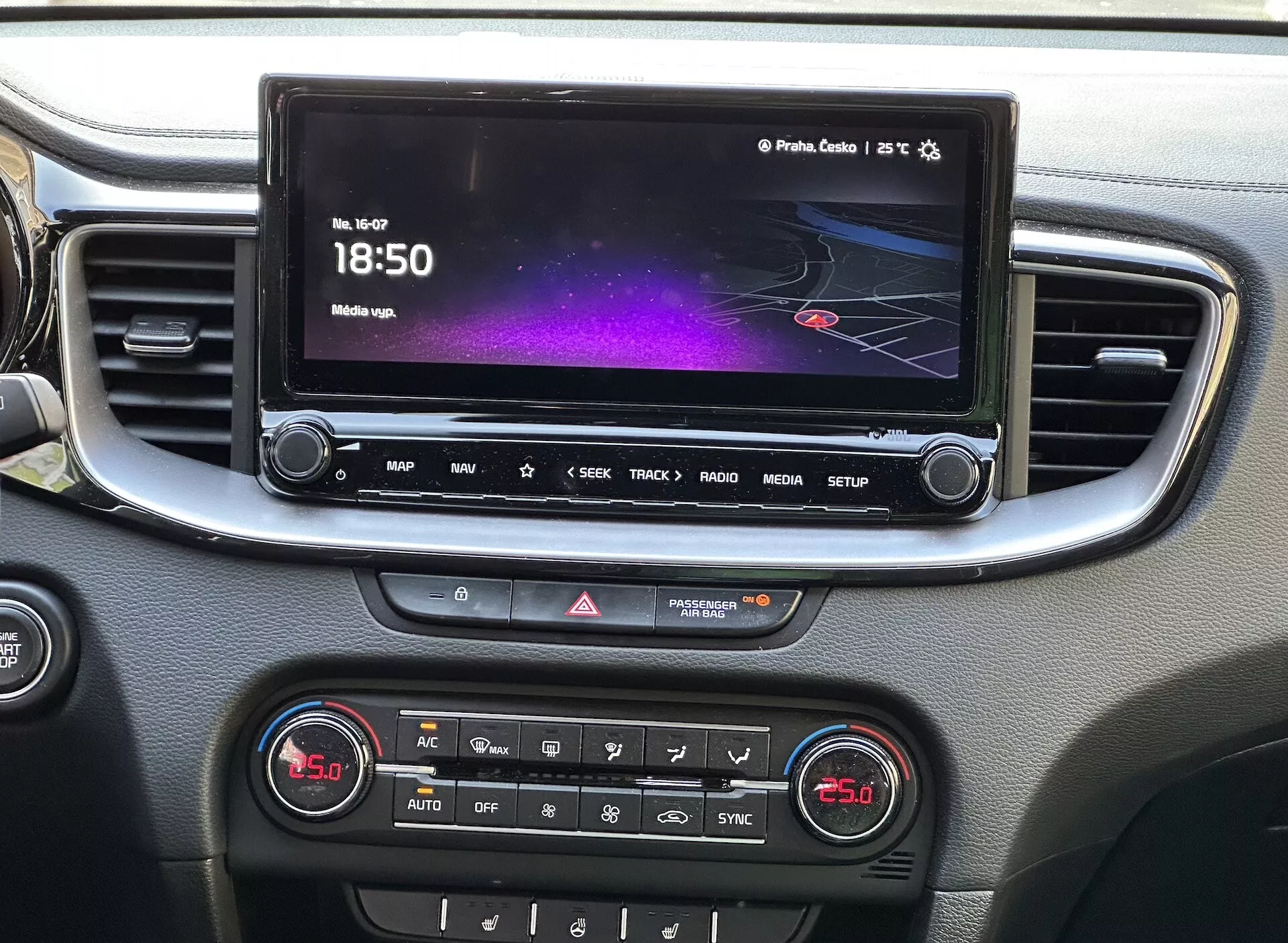 Vista detalhada do interior do Kia Xceed 2022 mostrando a consola central com um ecrã tátil de infotainment a exibir uma interface colorida com navegação e informação dos média, uma fila de botões de acesso rápido, controlo de luzes de emergência e um painel de controlo climático de zona dupla, tudo enquadrado com um acabamento a prata.