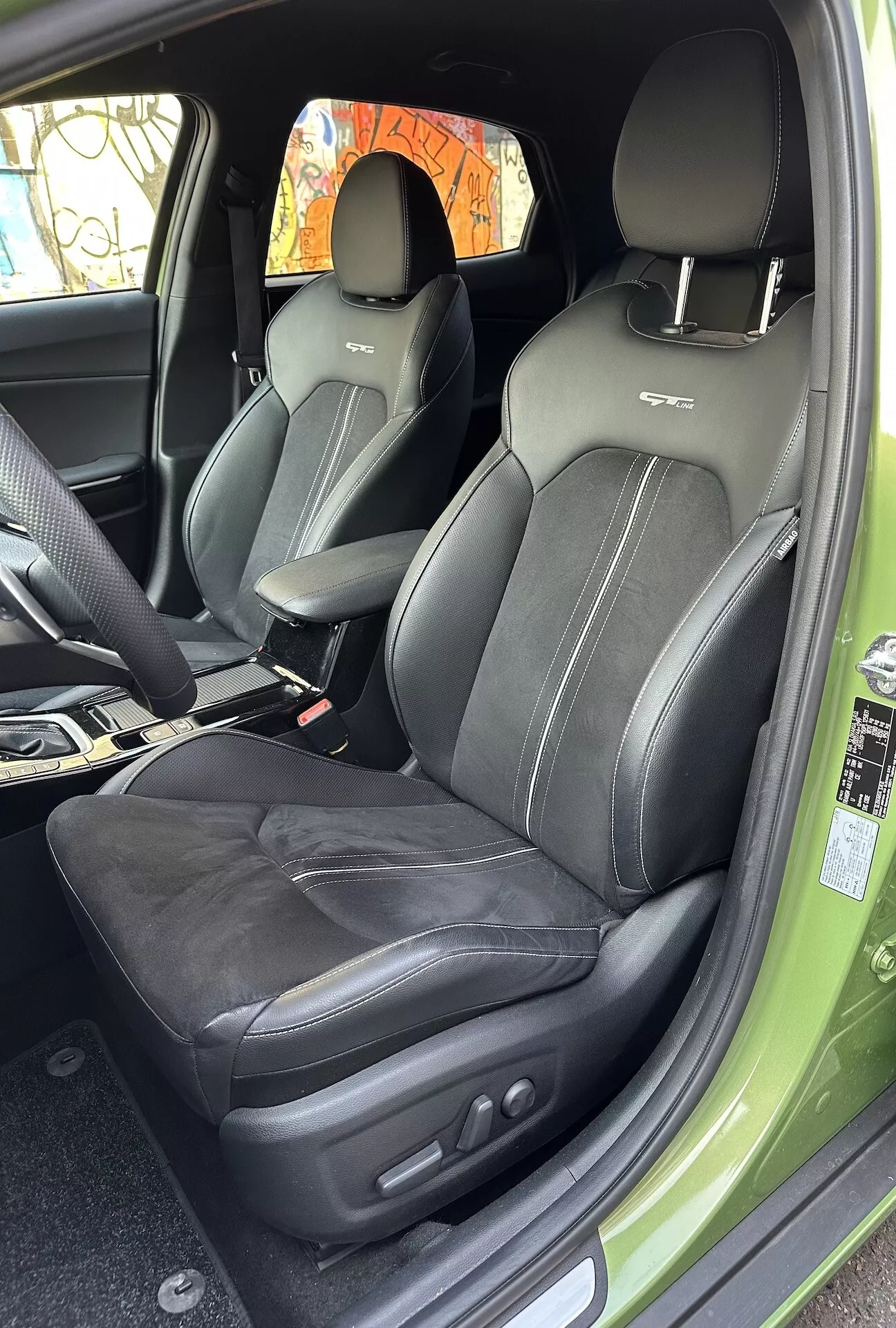 Vista interior do Kia XCeed 2022 focando no compartimento do passageiro da frente com estofamento em couro, costura intrincada e controles de ajuste do assento eletrônicos, de um ângulo de porta aberta mostrando um vislumbre do exterior verde vibrante.