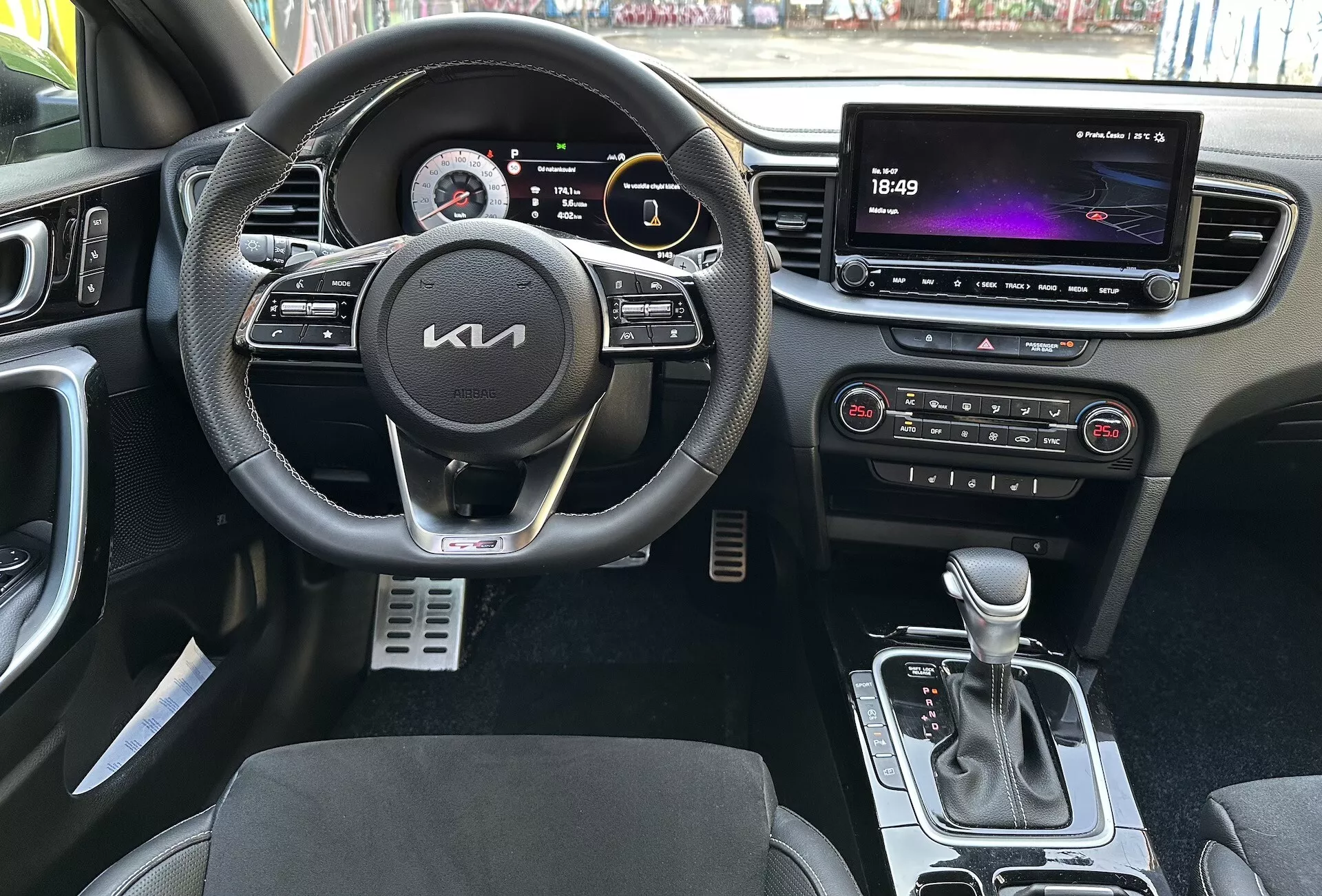 Vista interior do Kia XCeed 2022 destacando características voltadas para o motorista, como um volante multifuncional revestido a couro com botões para multimídia e controle de cruzeiro, um painel de instrumentos digital, um grande sistema de infotenimento touchscreen, controles automáticos de clima, alavanca de câmbio automática, bancos com estofamento bicolor e pedais esportivos de metal a partir de uma perspectiva de primeira pessoa.