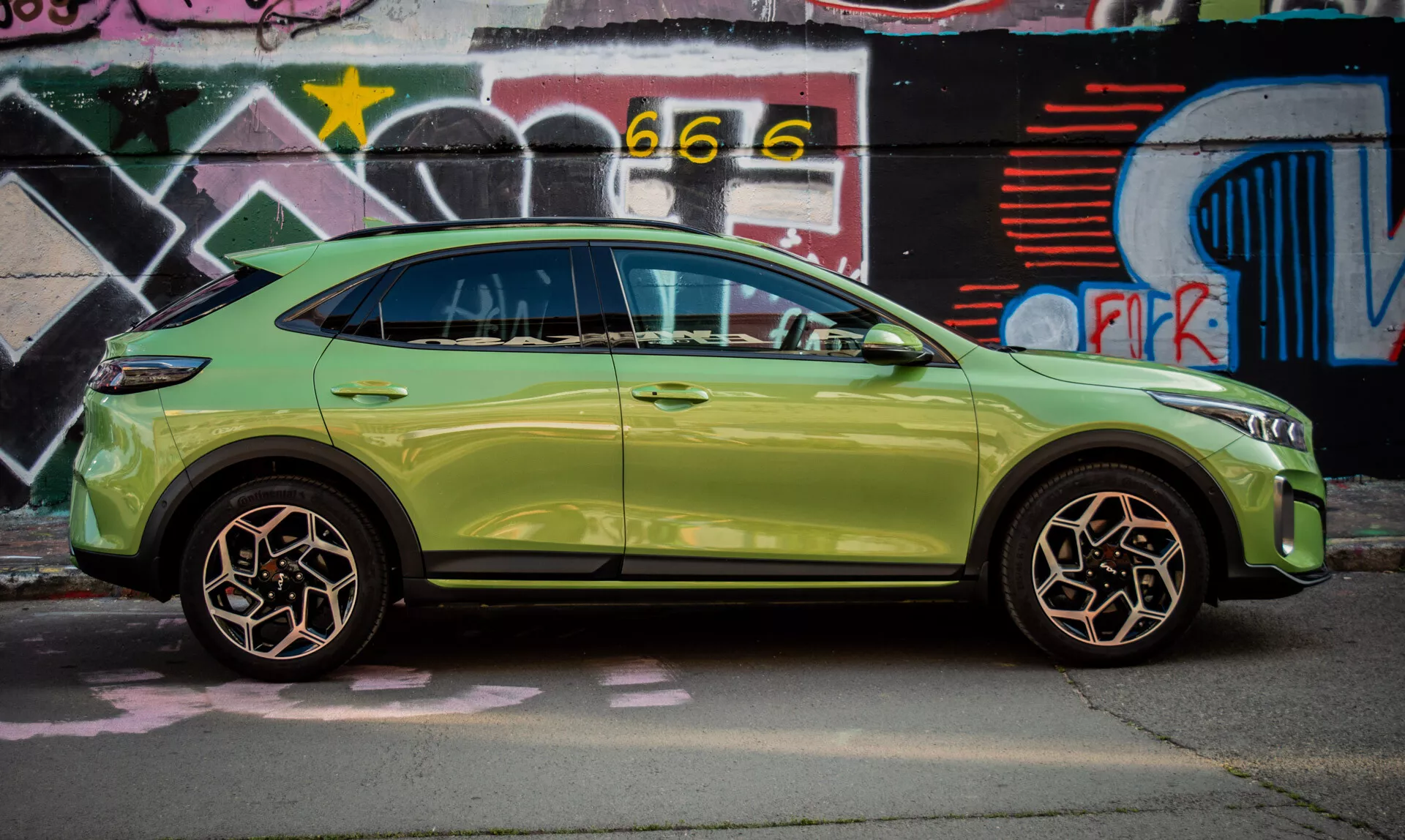 SUV compacto Kia XCeed de 2022 em verde-lima estacionado junto a uma parede coberta de grafite, exibindo seu design elegante e recursos avançados como entrada sem chave e sinais de direção integrados nos espelhos laterais.