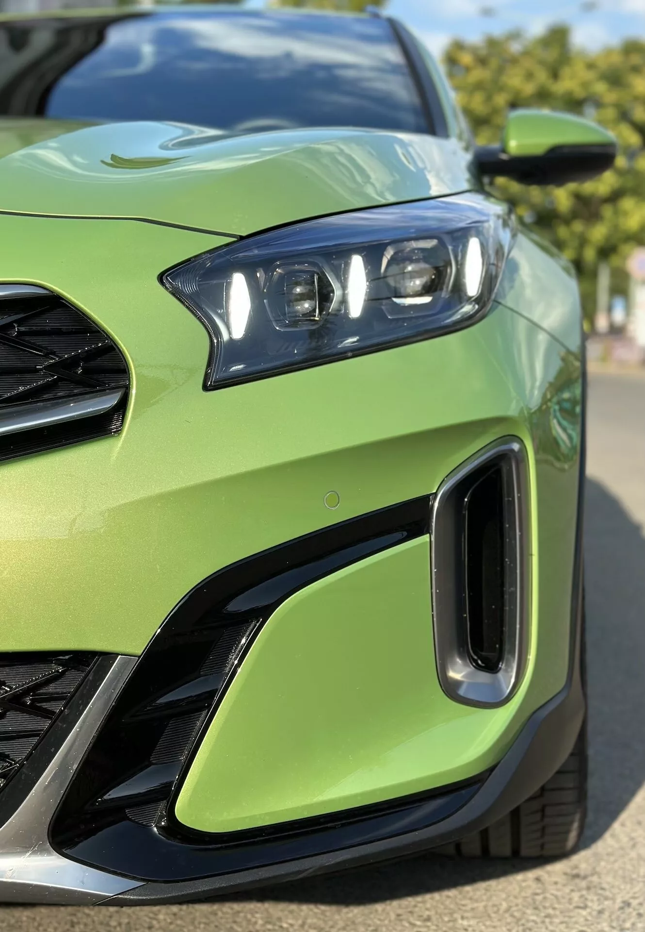Close-up do quarto frontal esquerdo da Kia Xceed 2022 mostrando em detalhe a montagem de farol LED, parte da grade, entradas de ar no para-choque dianteiro, contra um corpo de cor verde lima, enfatizando o design esportivo do veículo e tecnologia de iluminação avançada.