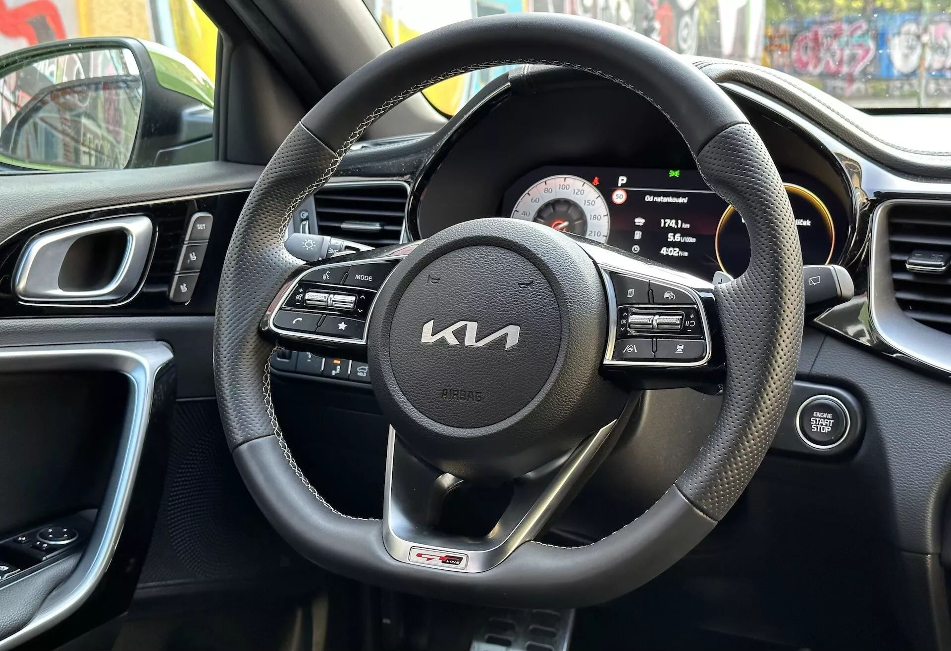 Vista interior do Kia XCeed 2022 pela perspectiva do motorista exibindo volante envolto em couro preto com controles multifuncionais e emblema da linha GT, cluster de instrumentos digital-analógico, parte do console central com ventilações, design ergonômico do painel de instrumentos, e painel da porta do motorista com controles, refletindo ambiente urbano