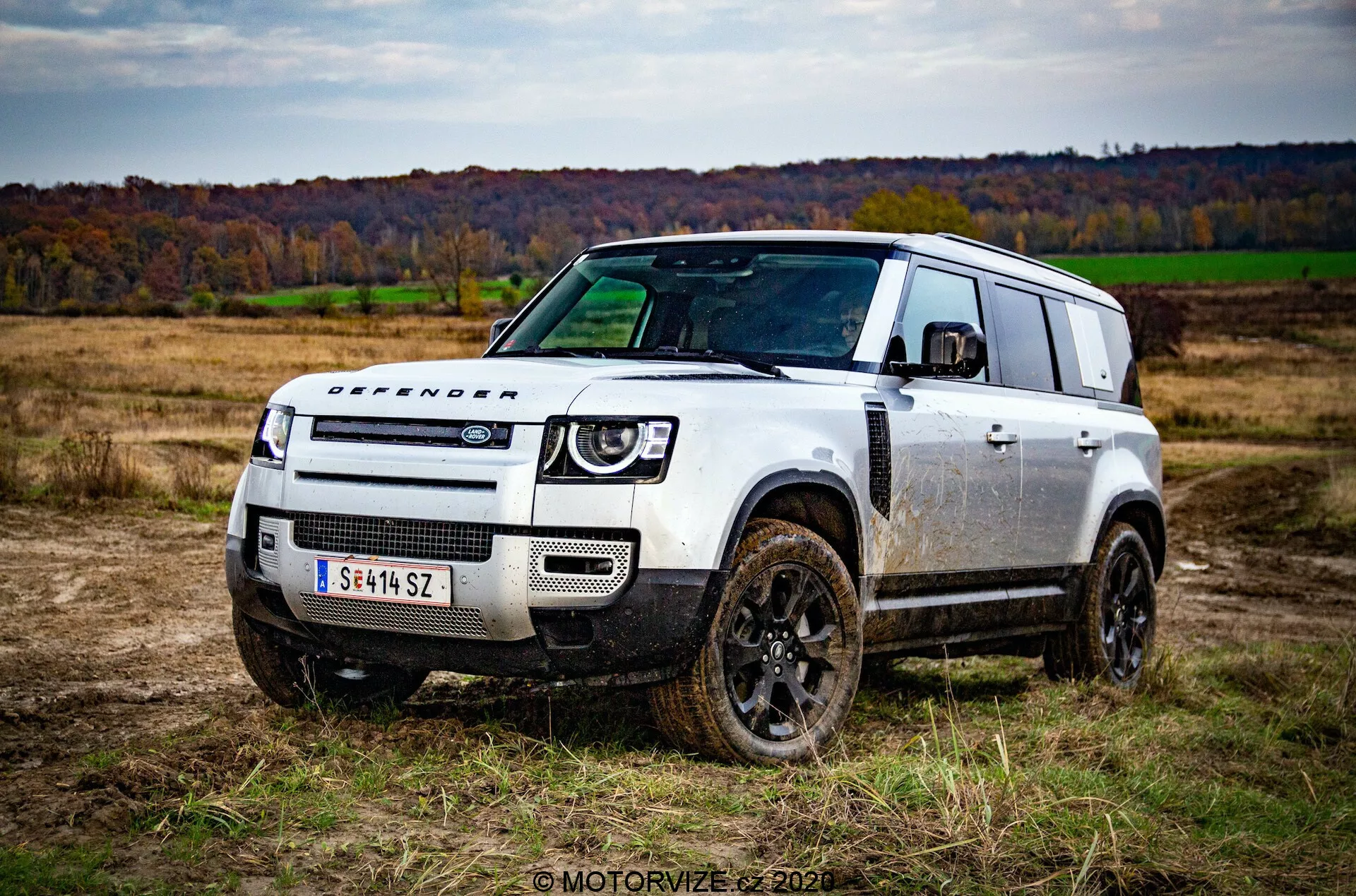 Imagem de um veículo off-road Land Rover Defender 2020 em um campo aberto com grama e árvores durante o outono, capturada em um ângulo de três quartos frontais. O design robusto e quadrado do veículo, coberto de manchas de lama, destaca suas capacidades off-road. Características notáveis incluem rodas escuras, letreiro 'DEFENDER' no capô, grade frontal minimalista com emblema Land Rover, faróis LED modernos e para-choques dianteiro robusto com placas de proteção. O design do teto escalonado com janelas alpinas e grandes janelas traseiras também adicionam à estética funcional do carro.