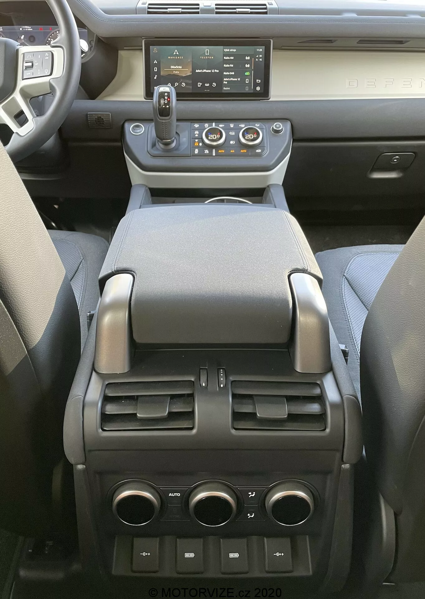 Vista interior do Land Rover Defender 2020 focando na consola central e na parte traseira da cabine da frente, exibindo um moderno sistema de infoentretenimento com tela sensível ao toque, saídas de ar centrais com botões de controle de clima, câmbio automático com opção de seleção manual, vários botões de controle, porta-copos, apoio de braço macio, compartimento de armazenamento, saídas de ar para controle de clima para os passageiros traseiros e portas USB e uma tomada de 12V na parte traseira, indicando uma combinação de tecnologia avançada, conforto e praticidade.