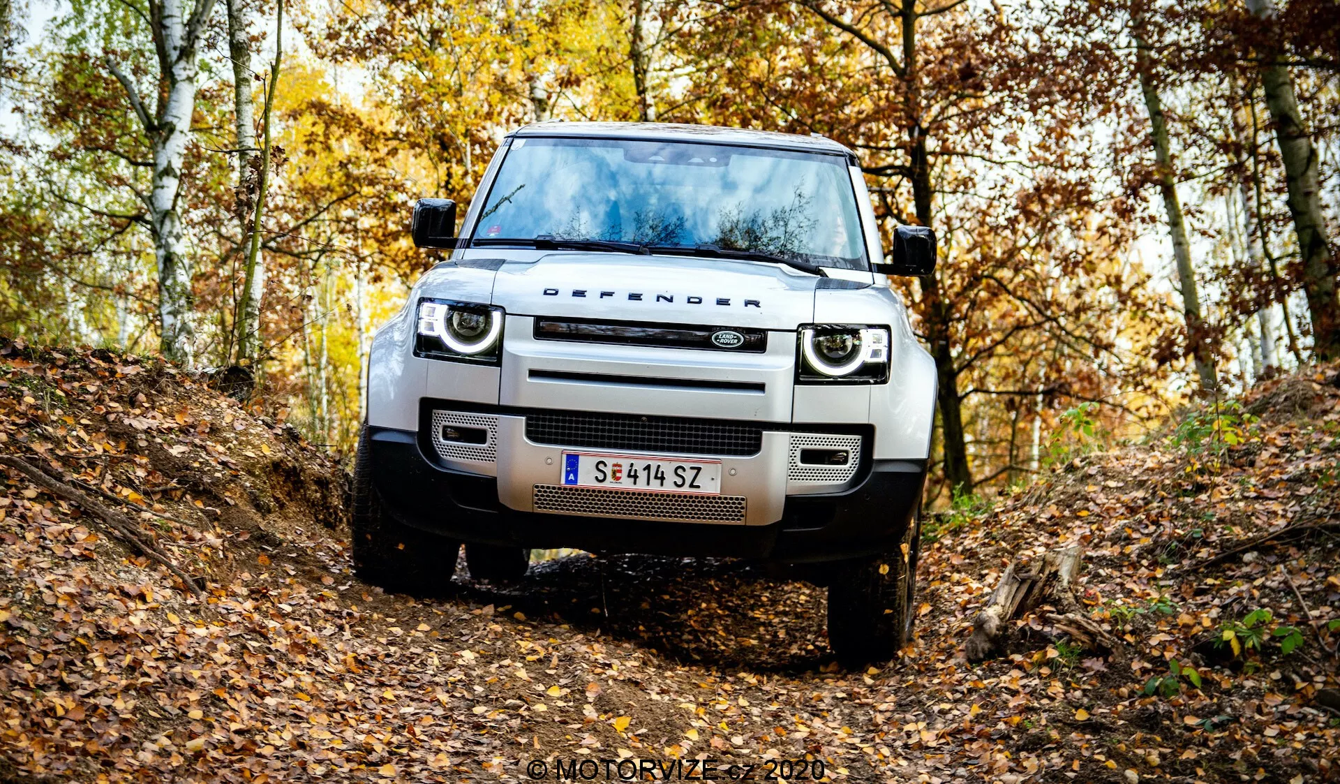 Vista frontal de um moderno Land Rover Defender em um cenário natural de floresta outonal em um caminho de terra irregular. A imagem captura a grade, faróis, capô, rodas e pneus do veículo, destacando sua altura do solo e capacidade off-road. Possui iluminação LED moderna, design simples da grade, sensores no para-choque dianteiro ou assistente de estacionamento, placa de aço e uma placa de matrícula austríaca. Folhas caídas e árvores com folhagem amarela e marrom criam um clima sazonal. Marca d'água 'MOTORYZE 2021' no canto inferior direito.
