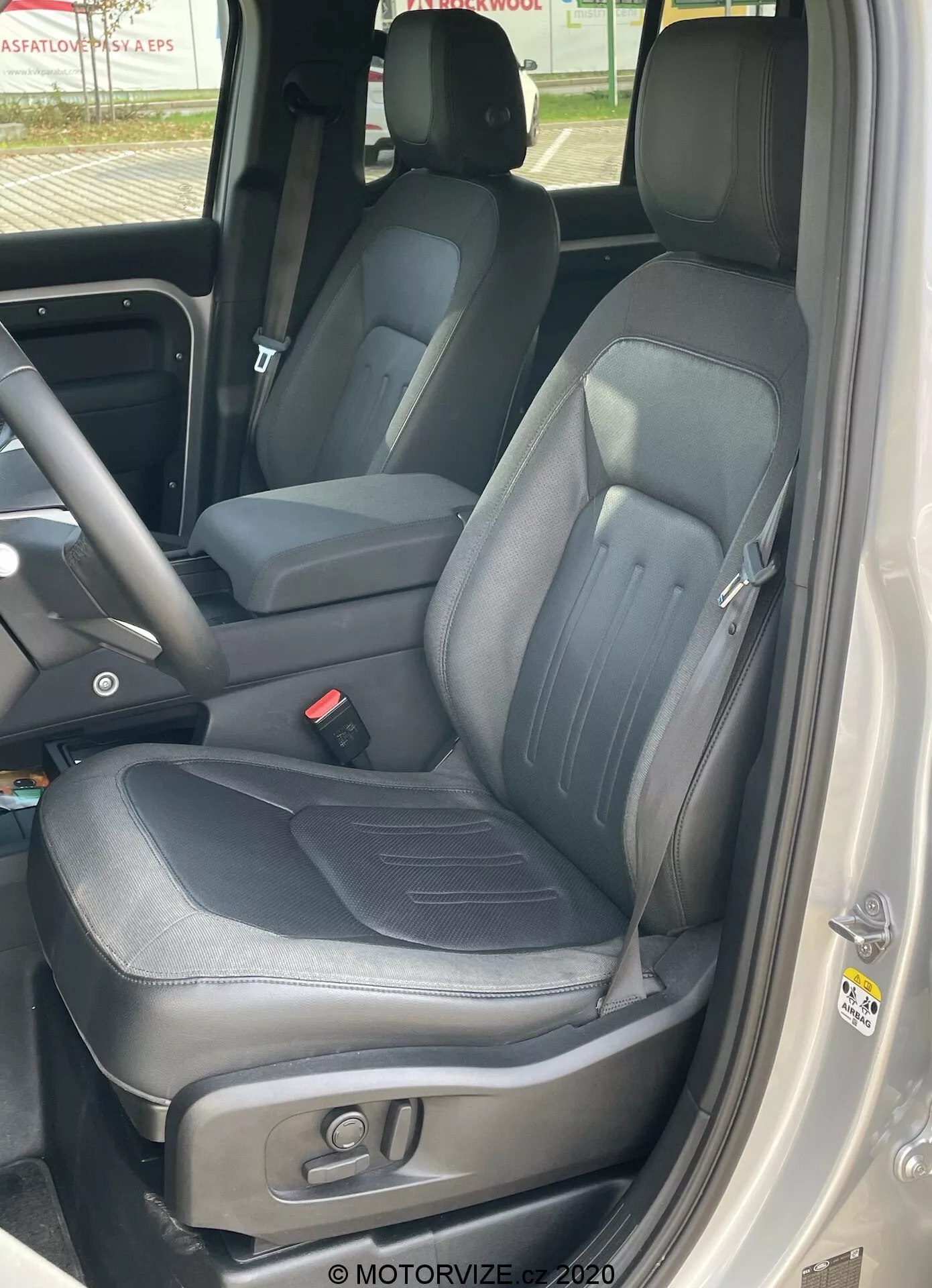 Vista aproximada do interior de um Land Rover Defender 2020 focando no banco do motorista, tirada de fora do carro com a porta aberta mostrando estofamento de couro preto com detalhes de costura visíveis, reforços laterais, encosto de cabeça integrado ajustável, controles eletrônicos para ajuste do assento, parte do console central indicando uma cabine dianteira espaçosa e o painel da porta com alça, botões de controle e alto-falantes.