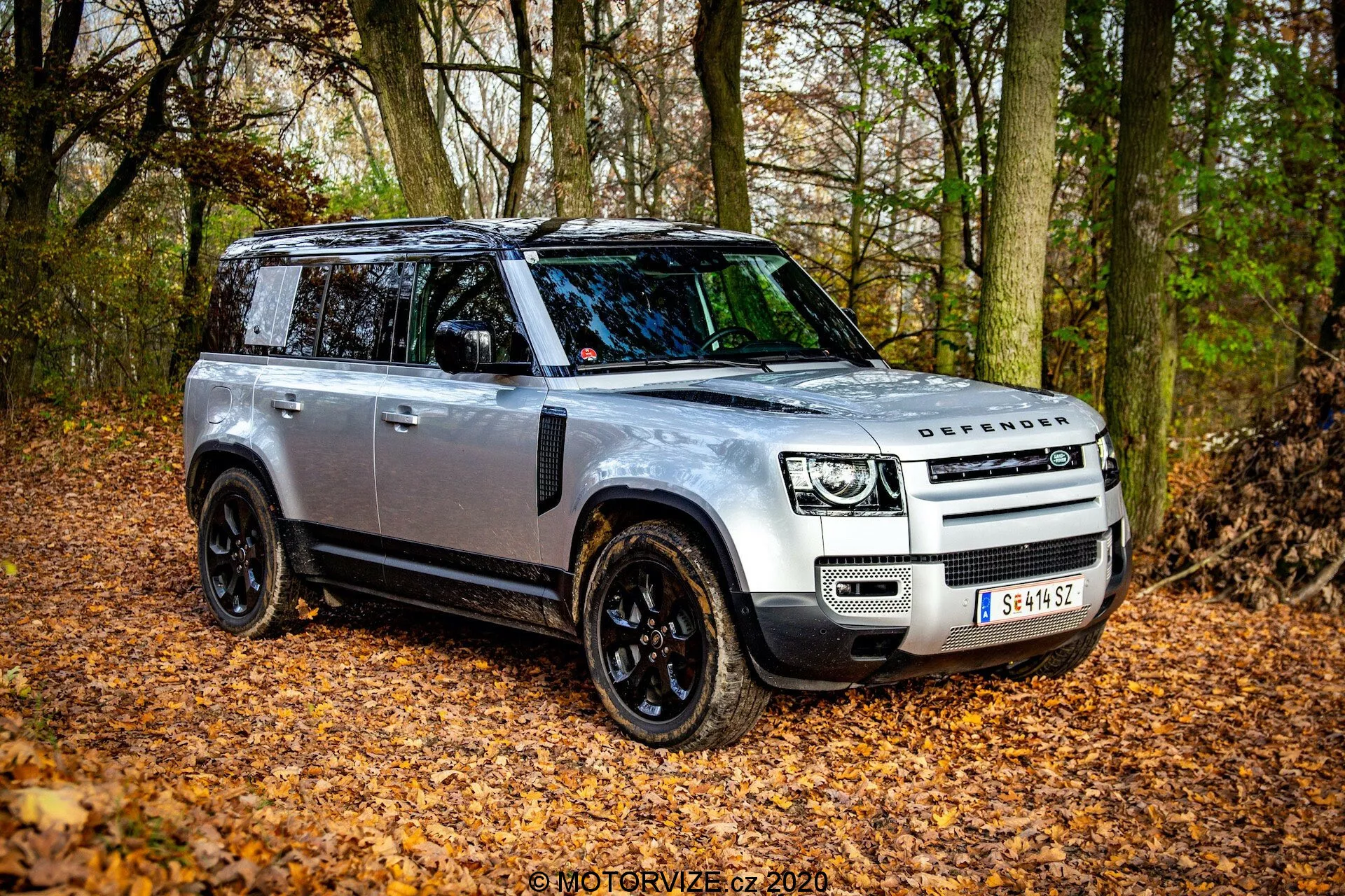 Land Rover Defender 2020 em ângulo frontal de três quartos, mostrando seu design robusto e características avançadas em um ambiente natural outonal com folhas caídas e árvores. O SUV tem uma carroceria prata com detalhes em preto, faróis de LED, teto panorâmico e rodas de liga leve pretas. A imagem capta o lado do motorista, destacando a grade frontal com o logotipo DEFENDER, espelhos laterais pretos com indicadores e possíveis placas de deslizamento para off-road.