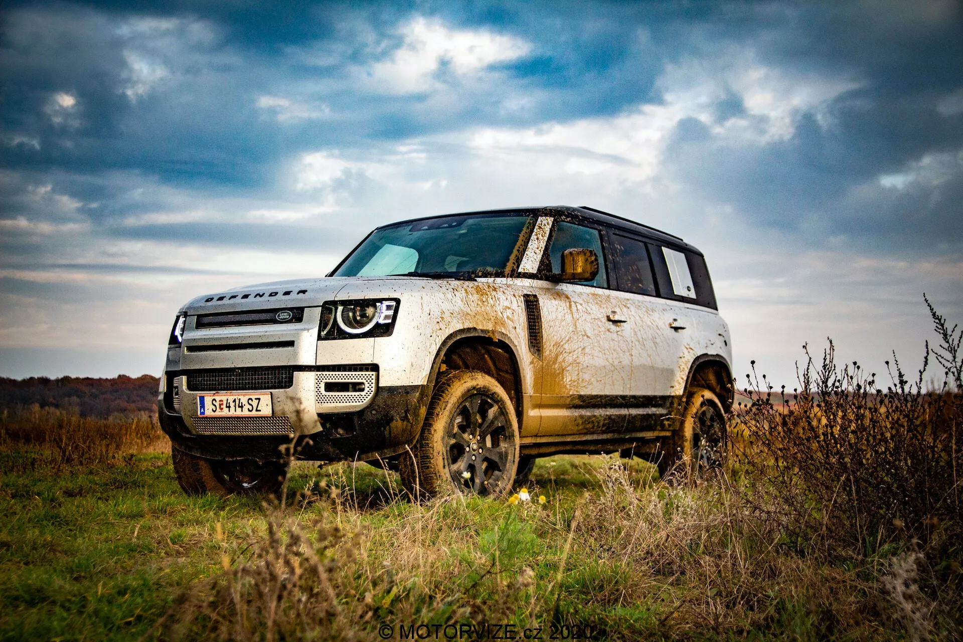 Imagem de um SUV Land Rover Defender 2020 ao ar livre, destacando sua capacidade off-road a partir de uma perspectiva de ângulo baixo. Enfatiza a altura do veículo, a robustez com os arcos das rodas, pneus e elementos protetores na parte inferior da carroceria. Captura uma vista frontal de três quartos, mostrando toda a frente e o lado até a porta traseira, acentuando elementos de design como o conjunto dos faróis, a grade e as linhas de contorno no capô. A lama e sujeira nos painéis laterais e nas rodas sugerem uso off-road, indicando durabilidade e capacidade off-road. Céus nublados e uma paisagem outonal ao fundo aumentam a sensação de aventura.