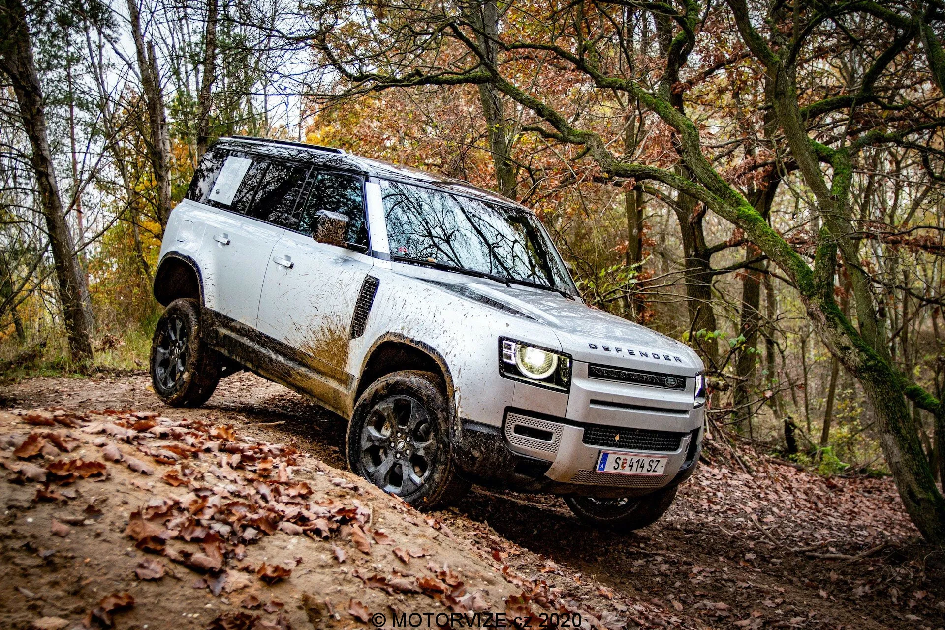 Perspectiva lateral de um SUV Land Rover Defender 2020 branco em ambiente off-road; design distinto com vista da grade e para-choques dianteiros, faróis LED modernos, rodas de liga preta com pneus off-road, espelhos retrovisores laterais implantados, silhueta quadrada mostrando sujeira, maçanetas embutidas, linha de teto plana e alta distância do solo. Veículo negociando uma inclinação, mostrando suas capacidades off-road.