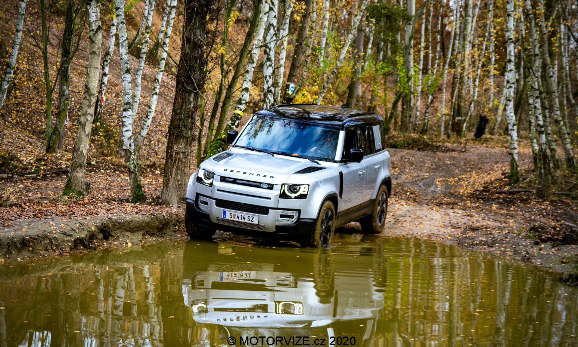 Imagem de um Land Rover Defender 2020 em um ambiente florestal, capturada a partir de um ângulo de três quartos frontal. O veículo demonstra capacidades off-road ao dirigir por um caminho lamacento cheio de água. O design apresenta faróis modernos, uma grade proeminente e uma placa de proteção. Trilhos de teto, pneus com sulcos profundos e uma superfície refletora de água adicionam um elemento estético. Cercado por bétulas e folhas caídas, a imagem captura a essência do outono.