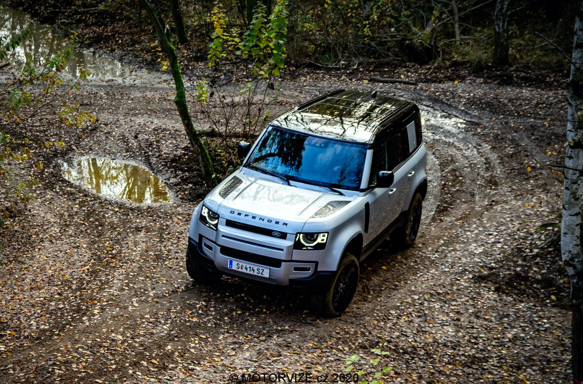 Vista aérea de um Land Rover Defender 2020 navegando por uma trilha off-road, com detalhes proeminentes como a grade frontal, capô, para-brisa e parte do lado do motorista, cercado por um ambiente natural com poças de água e lama, e árvores.