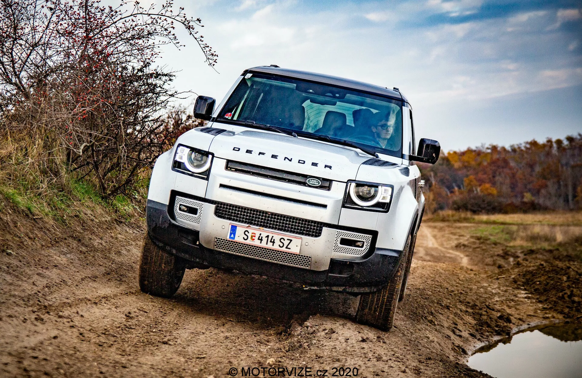 Vista de três quartos dianteira do Land Rover Defender 2020, SUV branco, quadrado e off-road com capô plano, arcos das rodas quadrados, luzes de condução diurna LED circulares, faróis adaptativos, grade em formato de colmeia com logotipo da marca, placa de proteção no para-choque dianteiro, postura robusta e elevada, espelho lateral com pisca integrado e ênfase na grade, faróis e para-choque dianteiro. Veículo em caminho não pavimentado com sujeira no exterior, rodeado por ambiente terroso e outonal. Foto profissional bem iluminada com fundo desfocado e marca de água 'MOTORVIZE © 2020'.
