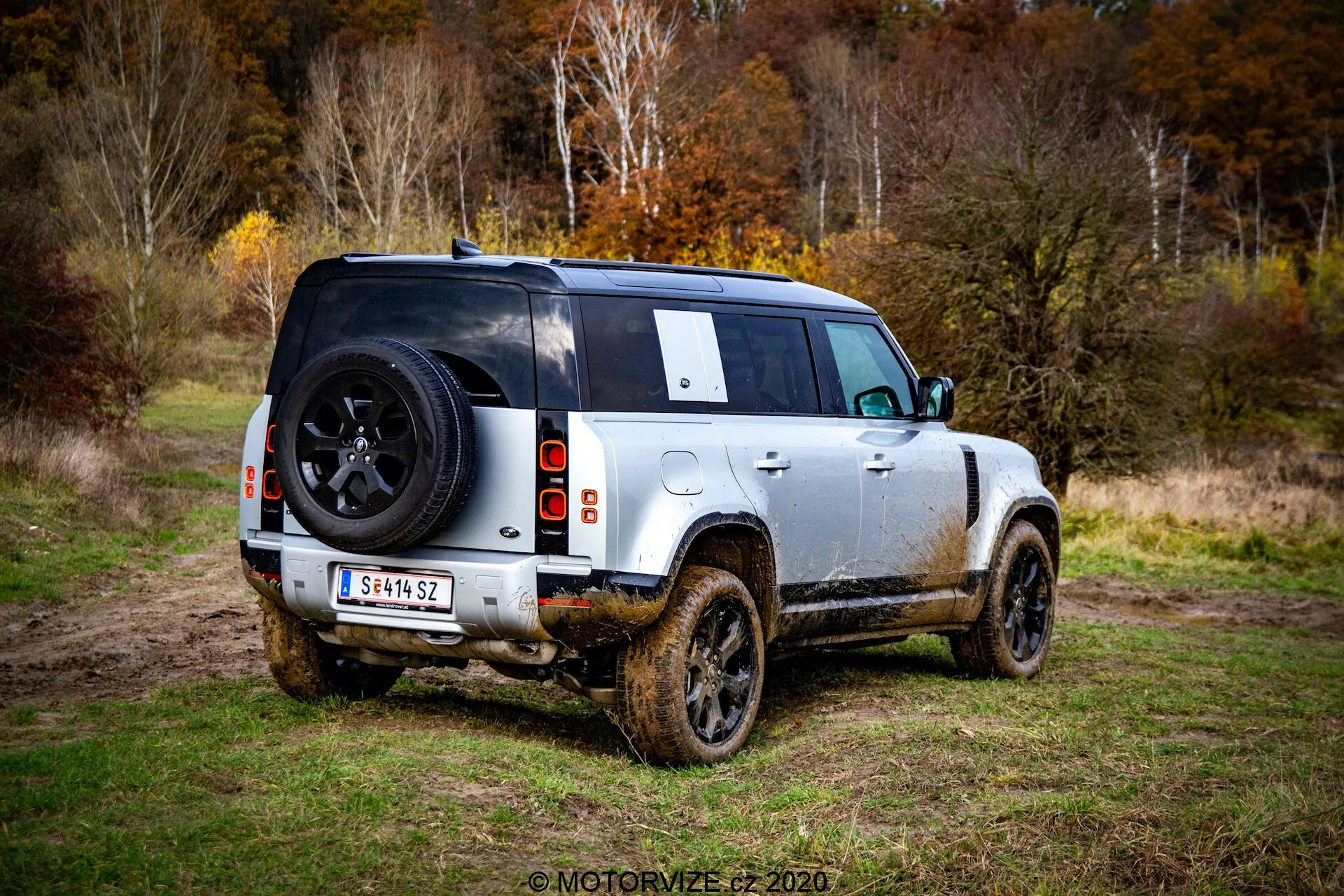 Vista traseira de três quartos de um Land Rover Defender 2020 contemporâneo, um SUV de quatro portas estacionado em uma área gramada com lama visível nas laterais, indicando uso recente fora de estrada. A foto tirada do nível do solo destaca os perfis traseiro e lateral, com foco no design traseiro incluindo uma luz traseira visível, roda traseira, área de carga e um pneu sobressalente montado na porta traseira com logotipo da marca. O veículo exibe um esquema de cores de dois tons com corpo claro e teto preto, rodas de liga leve com pneus robustos para todos os terrenos, alta distância ao solo e uma janela traseira parcialmente obstruída pela roda sobressalente e um limpador de para-brisa traseiro. Os lados e painéis inferiores enlameados sugerem que o SUV foi conduzido em terreno lamacento. O fundo sugere uma área de floresta ou matagal com luz suave, indicando um céu nublado ou condições de pouca luz.