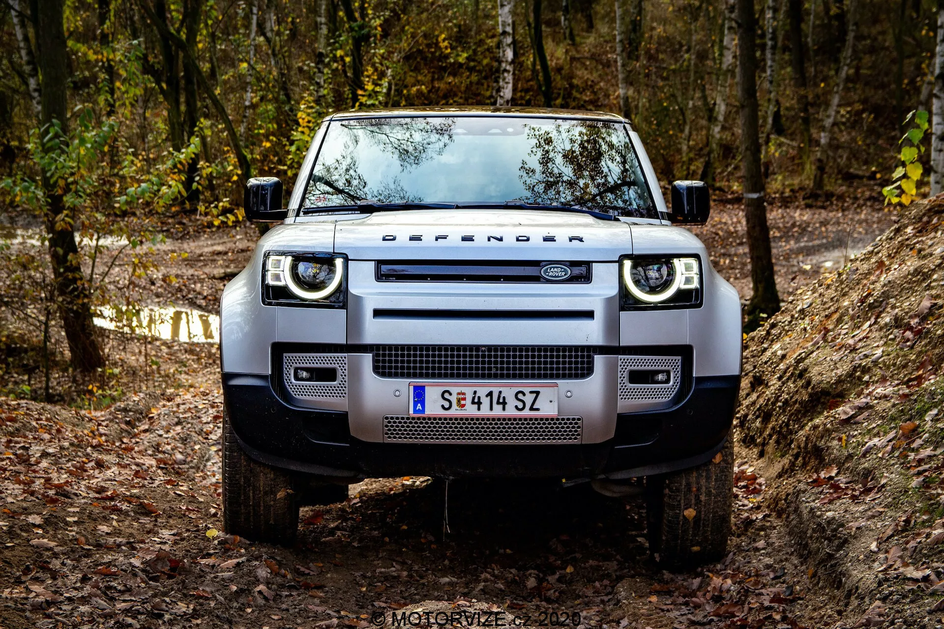 Vista frontal de um Land Rover Defender 2020 em um trilho off-road, mostrando sua presença robusta e dominante. Detalhes visíveis incluem a grade distintiva com a inscrição 'DEFENDER', faróis retangulares modernos com luzes diurnas de LED, para-choque dianteiro largo com faróis de neblina integrados, placas de proteção robustas, capô plano com bordas pronunciadas, pneus off-road robustos montados em rodas de liga leve e espelhos laterais funcionais com indicadores de direção integrados. O veículo está localizado em uma área arborizada com folhagem de outono, destacando seu espírito aventureiro e off-road.