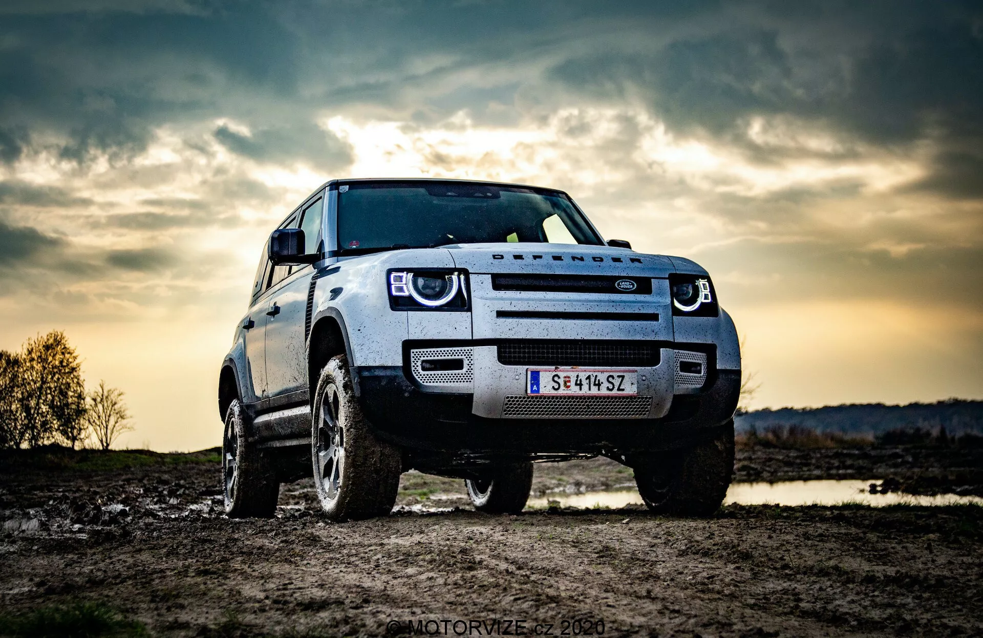 A imagem mostra um SUV Land Rover Defender 2020 estacionado ao ar livre, possivelmente em um ambiente off-road. Capturada de uma perspectiva de ângulo baixo, destaca a estatura robusta e altura do veículo. A vista frontal de três quartos mostra a grade dianteira, faróis, capô e para-lama e roda do lado do motorista. As luzes diurnas de LED distintas são visíveis, sugerindo características modernas de iluminação. A grade frontal ousada provavelmente reflete a identidade do fabricante, e uma câmera ou sensor frontal na grade indica sistemas avançados de assistência ao motorista. O céu dramático ao fundo, possivelmente com iluminação de fim de tarde ou crepúsculo, adiciona um senso de aventura, enquanto as rodas robustas e a alta distância do solo enfatizam a capacidade off-road.