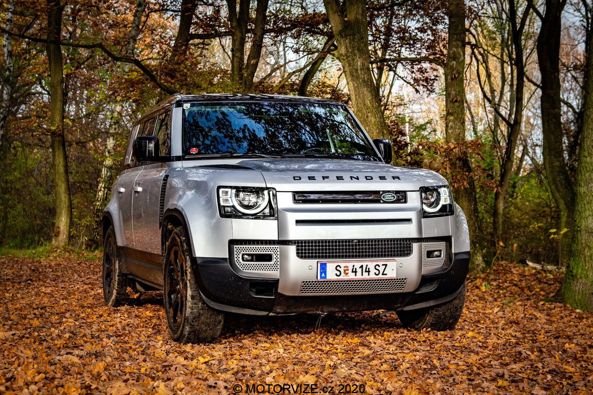Vista angular frontal de um Land Rover Defender 2020 moderno em um cenário off-road com folhas caídas, capturada de uma perspectiva de ângulo baixo. A imagem destaca a grade frontal com o texto 'DEFENDER' no capô, faróis de LED com luzes diurnas quadradas e sensores modernos integrados no para-choque dianteiro. Rodas cobertas de lama e suspensão pneumática ajustável indicam a capacidade off-road do veículo. O lado do motorista, a porta dianteira e parte da porta traseira estão visíveis, mostrando o design robusto do veículo com rodas parcialmente viradas.