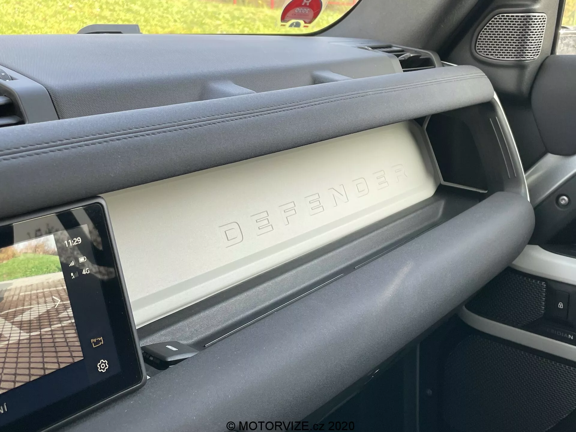 Interior do Land Rover Defender 2020 mostrando o lado do passageiro do painel de instrumentos a partir do ângulo do banco do motorista. A imagem captura o compartimento de luvas fechado com a palavra em relevo 'DEFENDER', exemplificando o design luxuoso e robusto do veículo. À direita, um moderno display touchscreen de infotainment mostra os controles do reprodutor de mídia e um mapa de navegação, indicando características tecnológicas avançadas. Os materiais do painel demonstram uma mistura de texturas premium e práticas, e a grelha do alto-falante é visível à esquerda, integrando-se com o design robusto do interior.