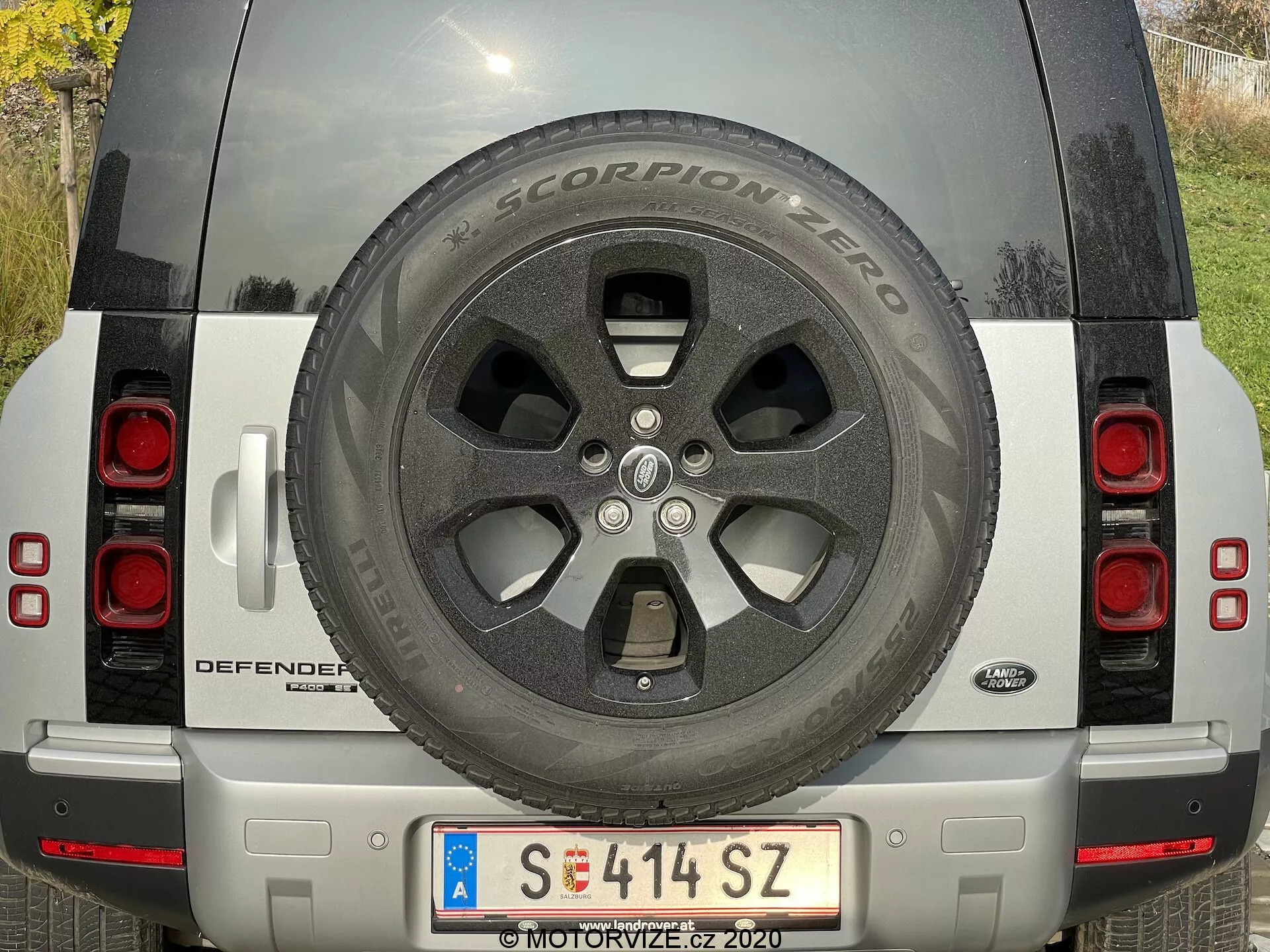 Vista traseira de um Land Rover Defender 2020 prateado com roda sobressalente visível, luzes traseiras, placa de matrícula, nome do modelo DEFENDER e emblema Land Rover, para-choque prateado com sensores e câmera e refletores. O SUV está estacionado à luz natural do dia, destacando o estado limpo e bem mantido do veículo.