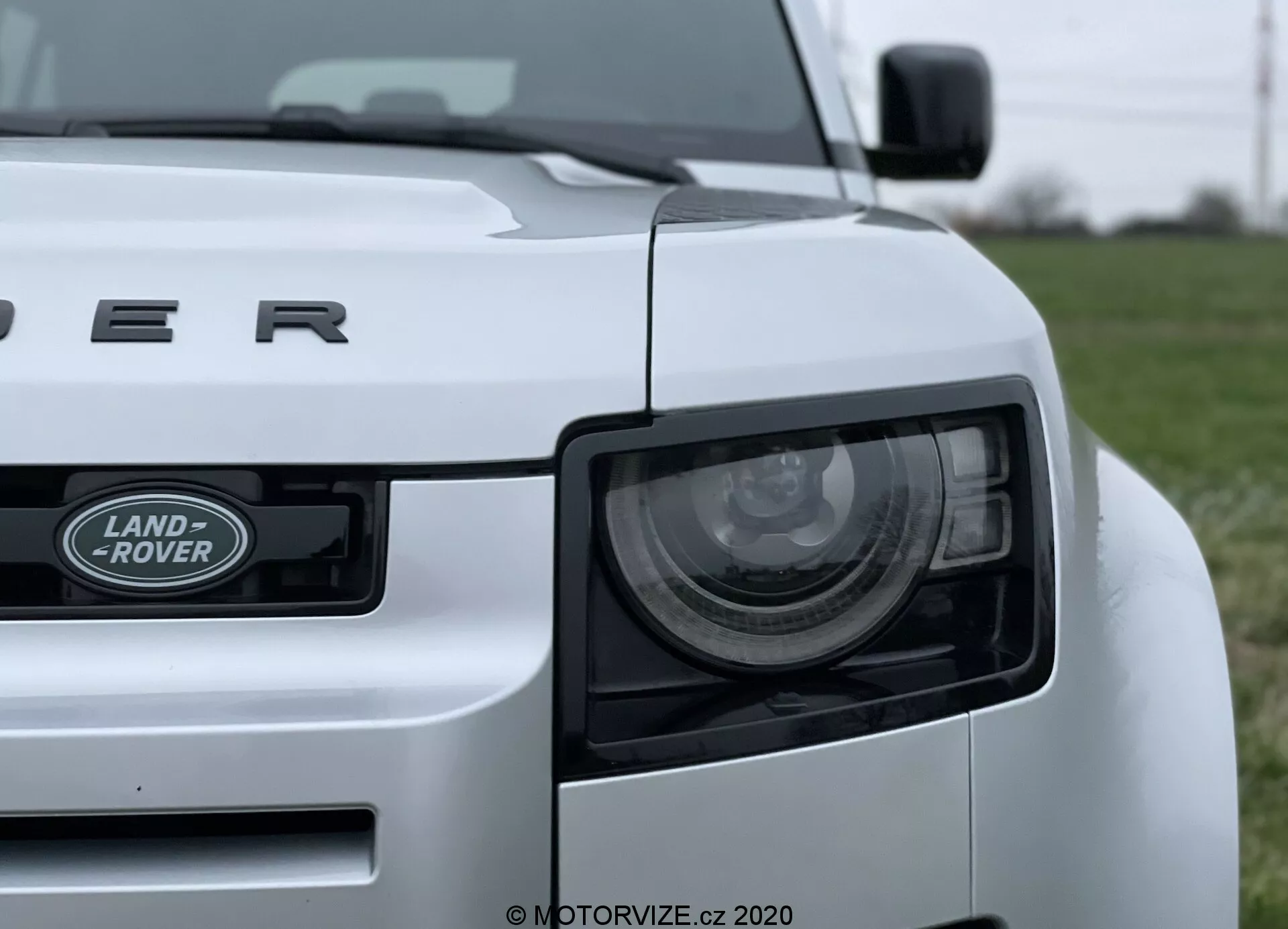 Imagem em close-up de um carro Land Rover Defender 2020 branco tirada de um ângulo baixo, mostrando o quarto dianteiro esquerdo do veículo. As características proeminentes incluem o farol, a grade com o emblema 'LAND ROVER' e o capô com a inscrição 'DEFENDER'. O farol possui elementos LED modernos, e o design robusto do carro é destacado por linhas e curvas visíveis. O carro está posicionado em um campo gramado, com o fundo desfocado para enfatizar o veículo.