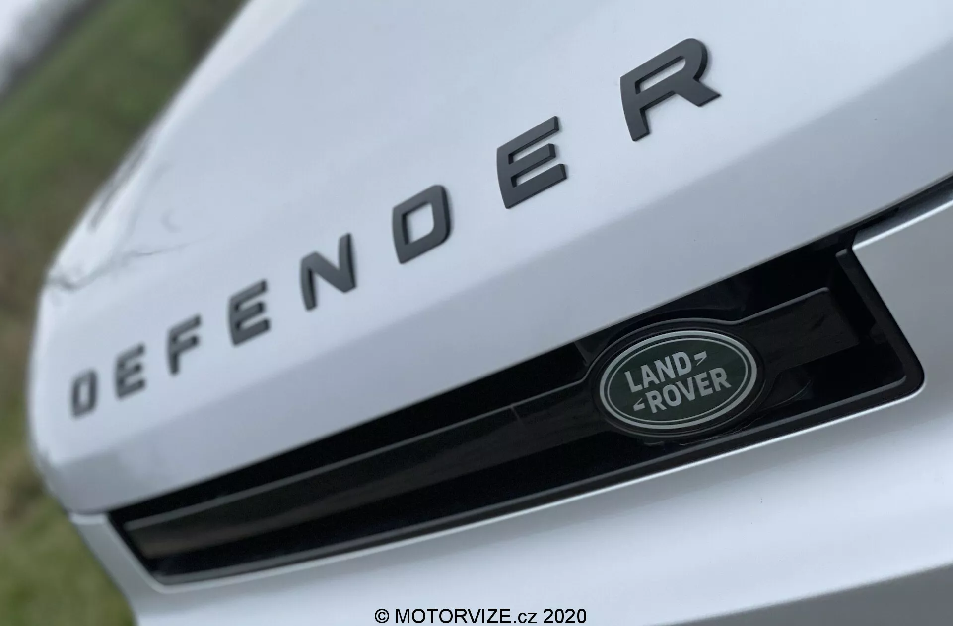 Vista aproximada da seção frontal de um Land Rover Defender 2020, com um foco destacado na placa de identificação 'DEFENDER' em letras prateadas em relevo na borda frontal do capô. Abaixo da placa de identificação, o emblema Land Rover em prata e verde com uma borda branca está centralmente posicionado na grade. A imagem é tirada de um ângulo baixo e ascendente com uma profundidade de campo rasa, destacando os elementos de marca, enquanto o fundo está desfocado.