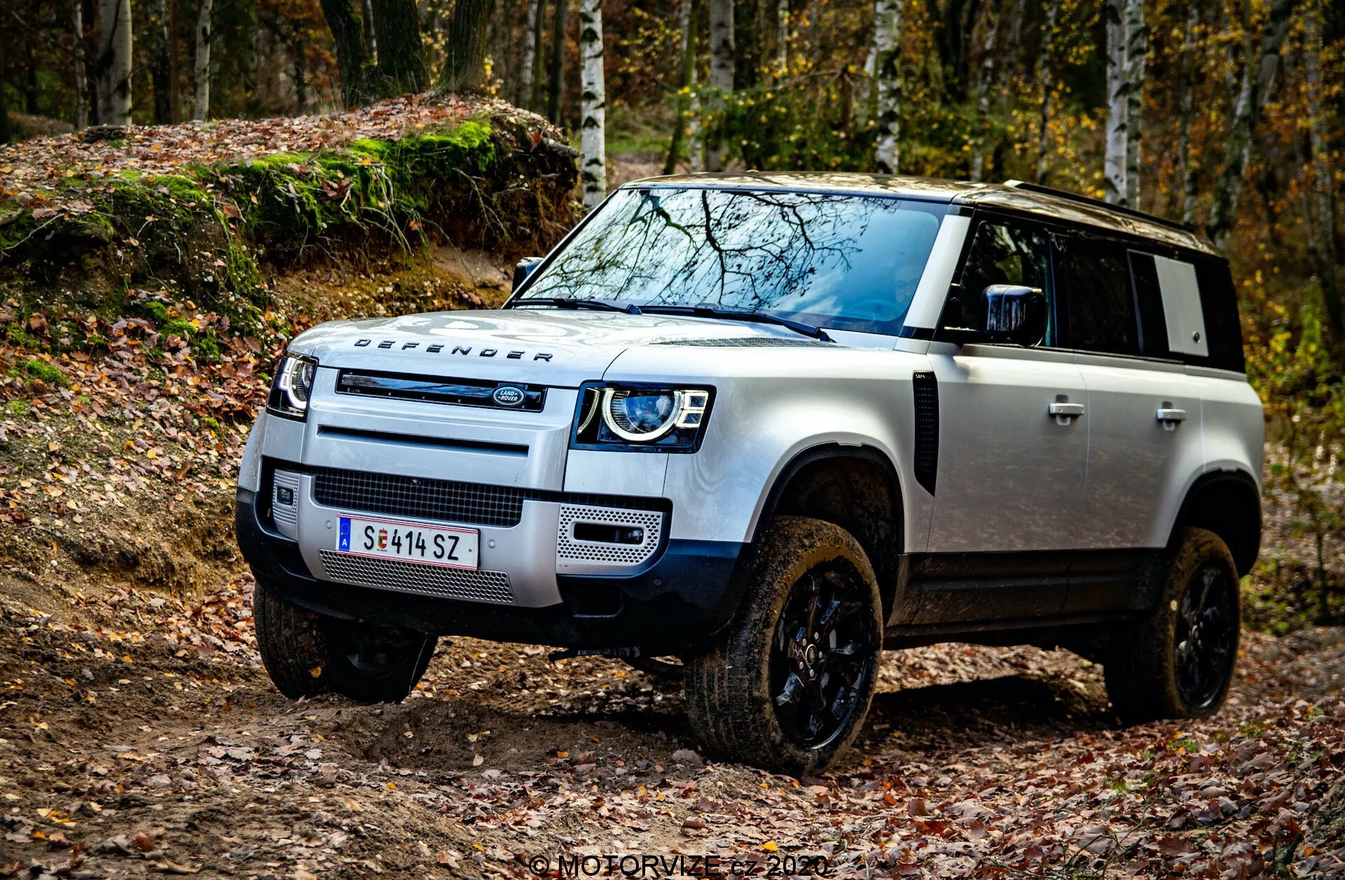 Fotografia em ângulo baixo de um Land Rover Defender 2020 capturada do quarto frontal esquerdo, destacando as características off-road robustas, como a grade frontal característica da Land Rover, faróis LED com luzes diurnas, placa de proteção no para-choque dianteiro, marca DEFENDER proeminente no capô, capô preto contrastante em corpo branco, entradas de ar elevadas para travessia de águas profundas, grandes arcos de roda com rodas off-road pretas, luzes de sinalização nos espelhos laterais e tecnologia avançada de assistência ao motorista perto do para-choque dianteiro. A imagem está ambientada em um ambiente off-road com folhas caídas, enfatizando a estética pronta para a aventura do veículo. Marca d'água 'MOTORVIZA 2021'.