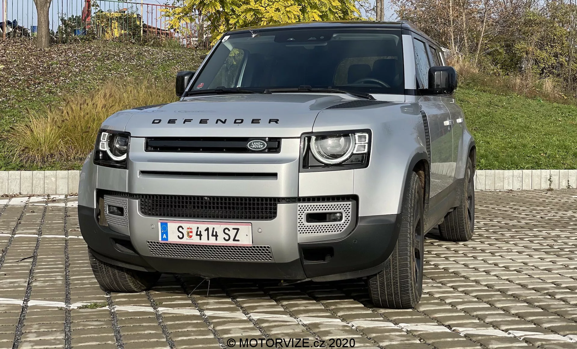 Vista frontal de um Land Rover Defender do modelo 2020, destacando seus faróis de LED de dois níveis distintivos, grade proeminente com logotipo Land Rover, pára-choque geométrico com placa de proteção prateada e outras características robustas que sugerem capacidades off-road. Também são visíveis o capô com a placa 'Defender', as entradas de ar e vistas parciais das rodas de cinco raios. O veículo é de uma cor neutra clara, provavelmente prata ou cinza.