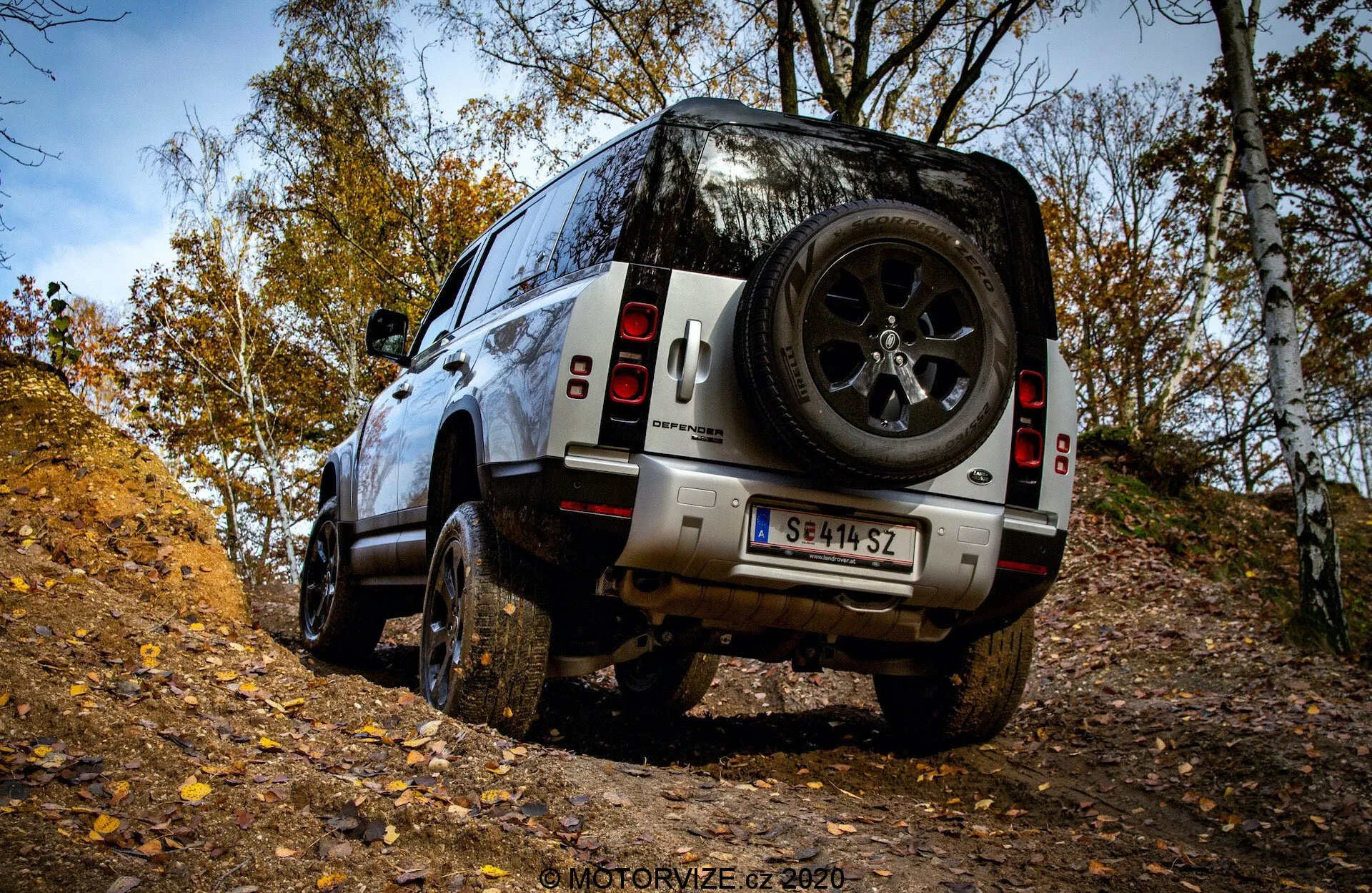 Vista traseira de três quartos do SUV Land Rover Defender 2020 em terreno off-road, destacando pneus robustos, ampla distância ao solo e roda de reserva traseira em um cenário de floresta no outono.