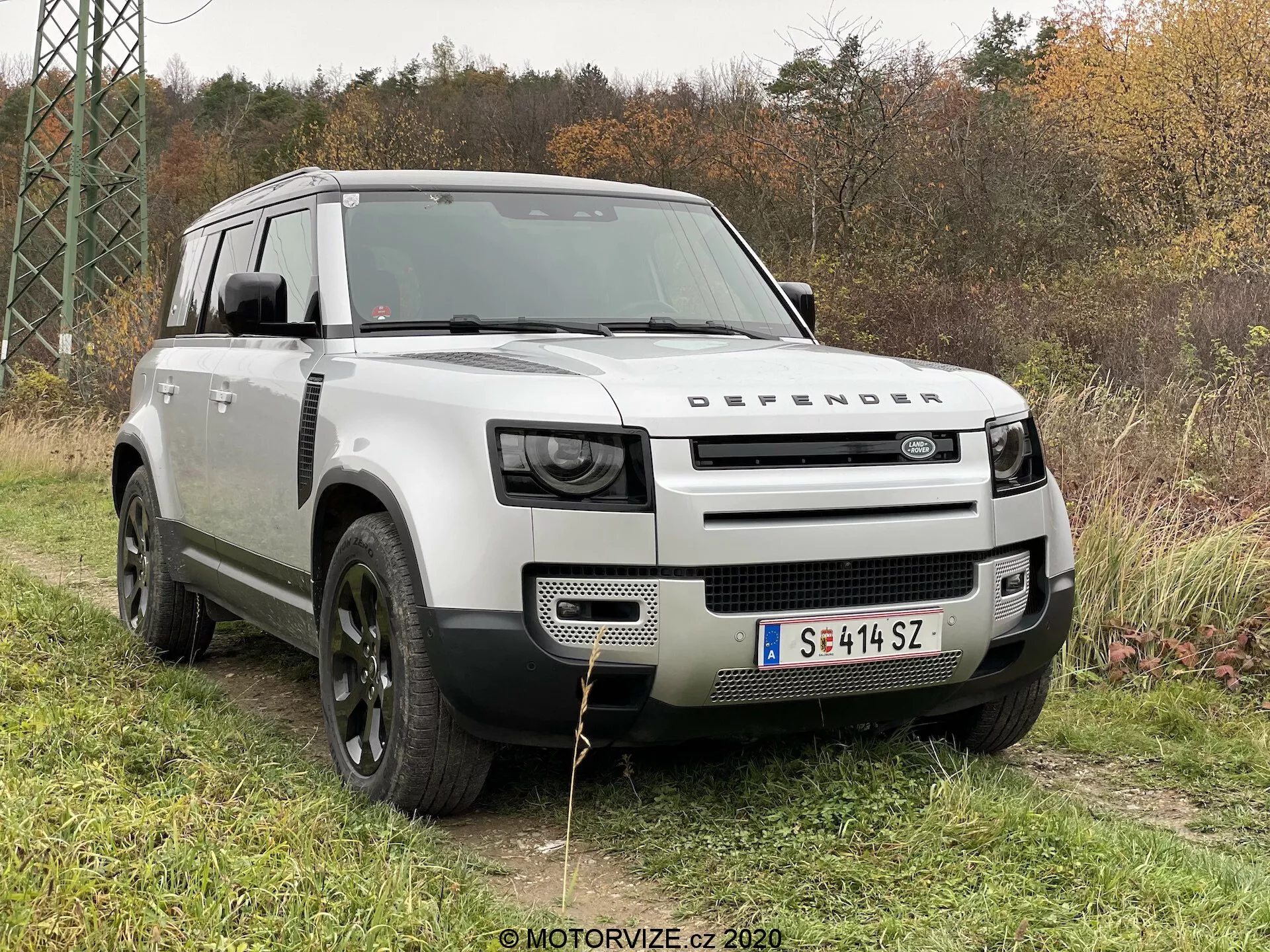 Esta imagem apresenta um Land Rover Defender 2020 em um cenário ao ar livre sobre a grama com folhagem e árvores ao fundo. Capturada de um ângulo frontal de três quartos, a foto destaca a grade proeminente com a marca 'DEFENDER', faróis LED modernos, capô robusto com design de concha, grandes rodas de liga preta com pneus off-road e uma carroceria prata claro que contrasta com os arcos das rodas e o teto pretos. Elementos de design como a placa de proteção frontal, janelas de admissão de ar 'alpine' e a estrutura quadrada enfatizam suas capacidades off-road e utilitárias.