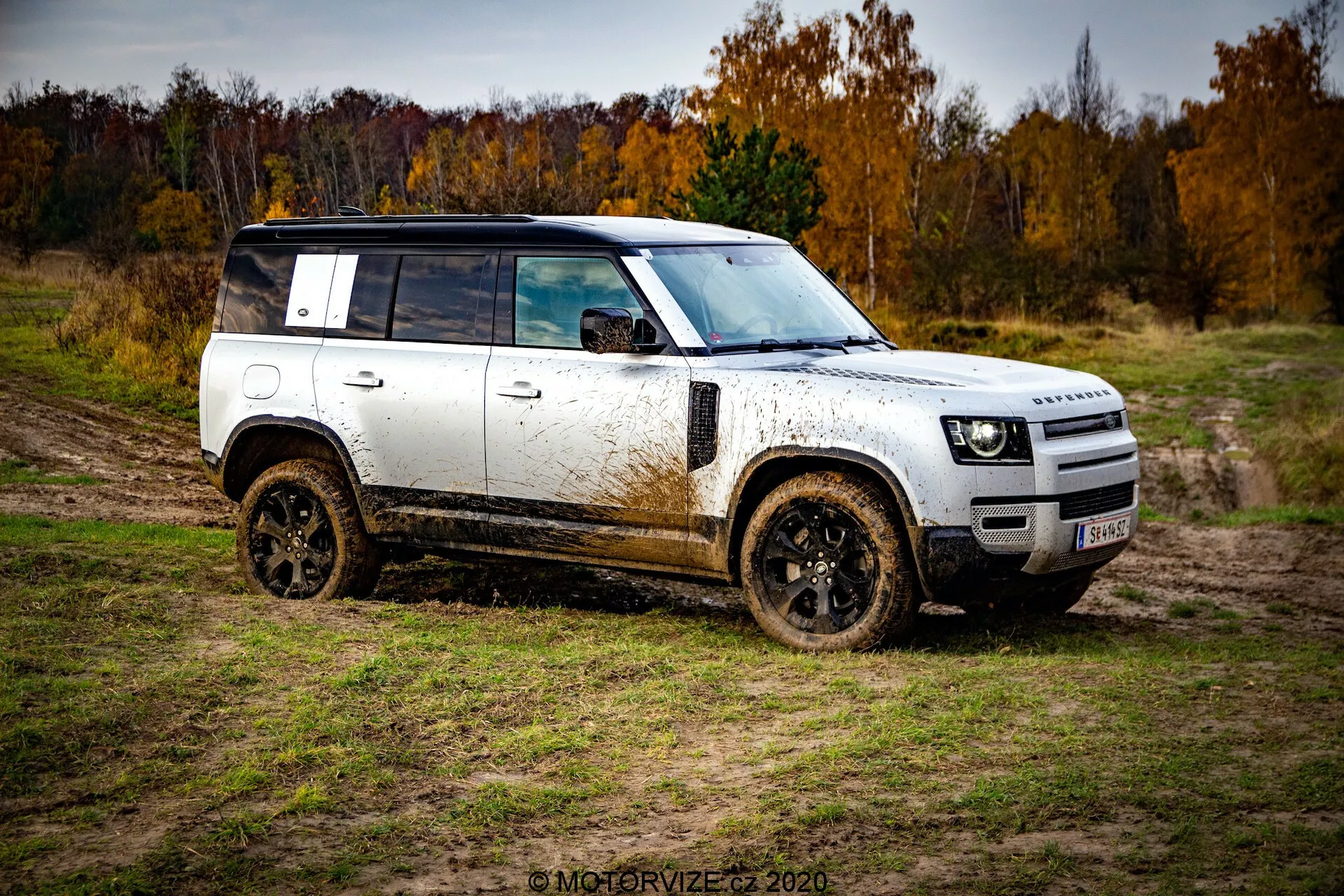 Perfil lateral de um SUV off-road Land Rover Defender 2020 mostrado em um ângulo de três quartos, exibindo sua carroceria branca com teto e rodas pretas, coberto de lama. Veículo posicionado diagonalmente do canto inferior esquerdo ao superior direito contra um terreno natural lamacento sob céu nublado com árvores outonais ao fundo.