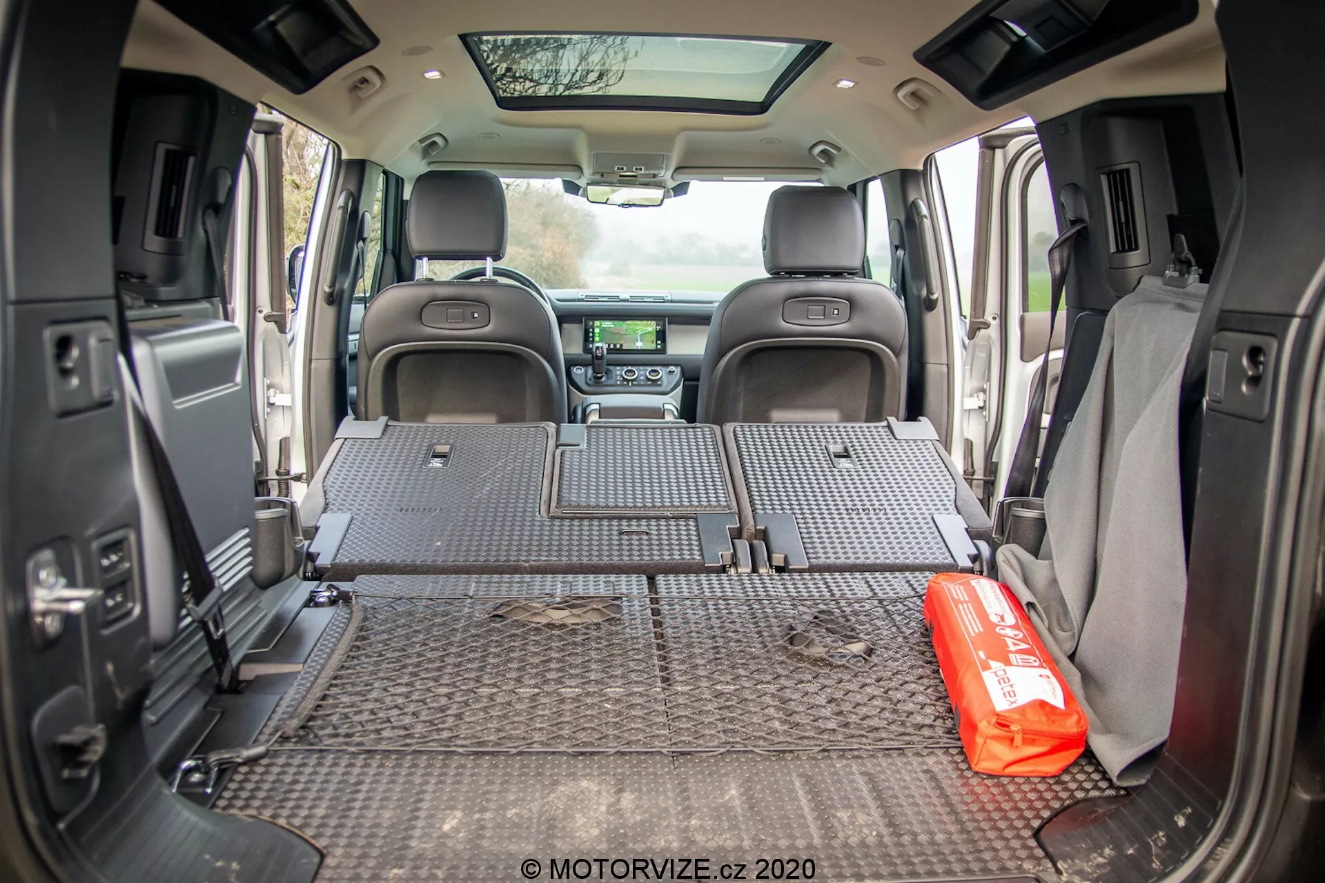 Vista interna do espaço de carga do Land Rover Defender 2020 tirada de um ângulo voltado para trás, mostrando o tapete protetor com padrões de placa diamante cobrindo todo o piso da área de carga. Os assentos traseiros estão rebatidos, revelando um espaço de carga espaçoso. As paredes laterais possuem acessórios como alto-falantes e porta-copos, sugerindo um design equilibrado entre funcionalidade e conforto. O painel frontal à distância é parcialmente visível, abrigando o sistema de infotainment e os controles de clima. Um kit de emergência vermelho está colocado de maneira proeminente no piso do espaço de carga, sugerindo o uso do veículo voltado para a família ou aventuras. A porta traseira aberta permite que a luz natural entre no espaço de carga.
