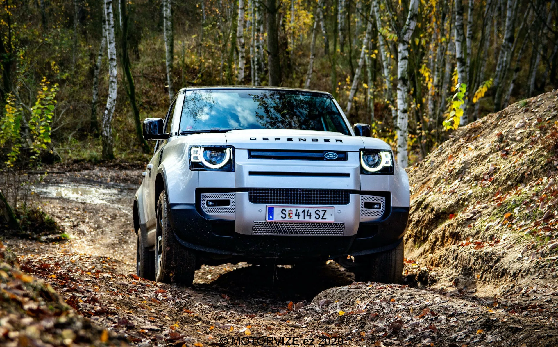 Vista frontal de um Land Rover Defender 2020, capturada de um ângulo baixo que enfatiza a grade, os faróis e o para-choque dianteiro. O SUV está posicionado em uma estrada não pavimentada com uma floresta ao fundo, destacando sua prontidão para off-road. Os elementos visíveis incluem o design proeminente 'DEFENDER', faróis de LED, para-lamas largos e pneus robustos.