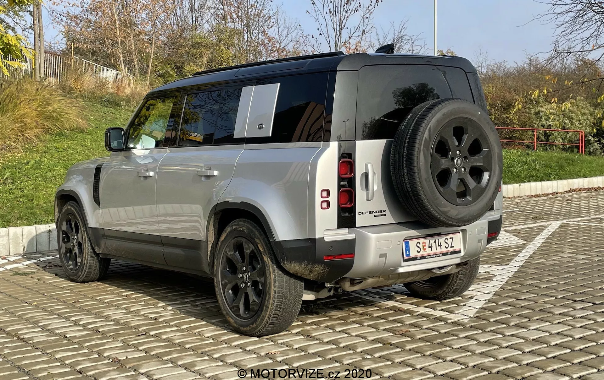 A imagem exibe a vista traseira de três quartos de um SUV Land Rover Defender 2020. Este ângulo permite uma visão clara do design robusto e quadrado do veículo, a pintura metálica prateada e as luzes traseiras de LED segmentadas verticalmente. Detalhes notáveis incluem um pneu sobressalente montado na porta traseira, para-choque traseiro robusto com sensores de estacionamento, grandes rodas de liga preta com paredes laterais grossas para uso off-road e vidros escuros nas janelas traseiras. Além disso, uma antena é visível no teto, sugerindo a presença de sistemas avançados de comunicação. O carro está estacionado em uma pavimentação intertravada sob um céu claro.