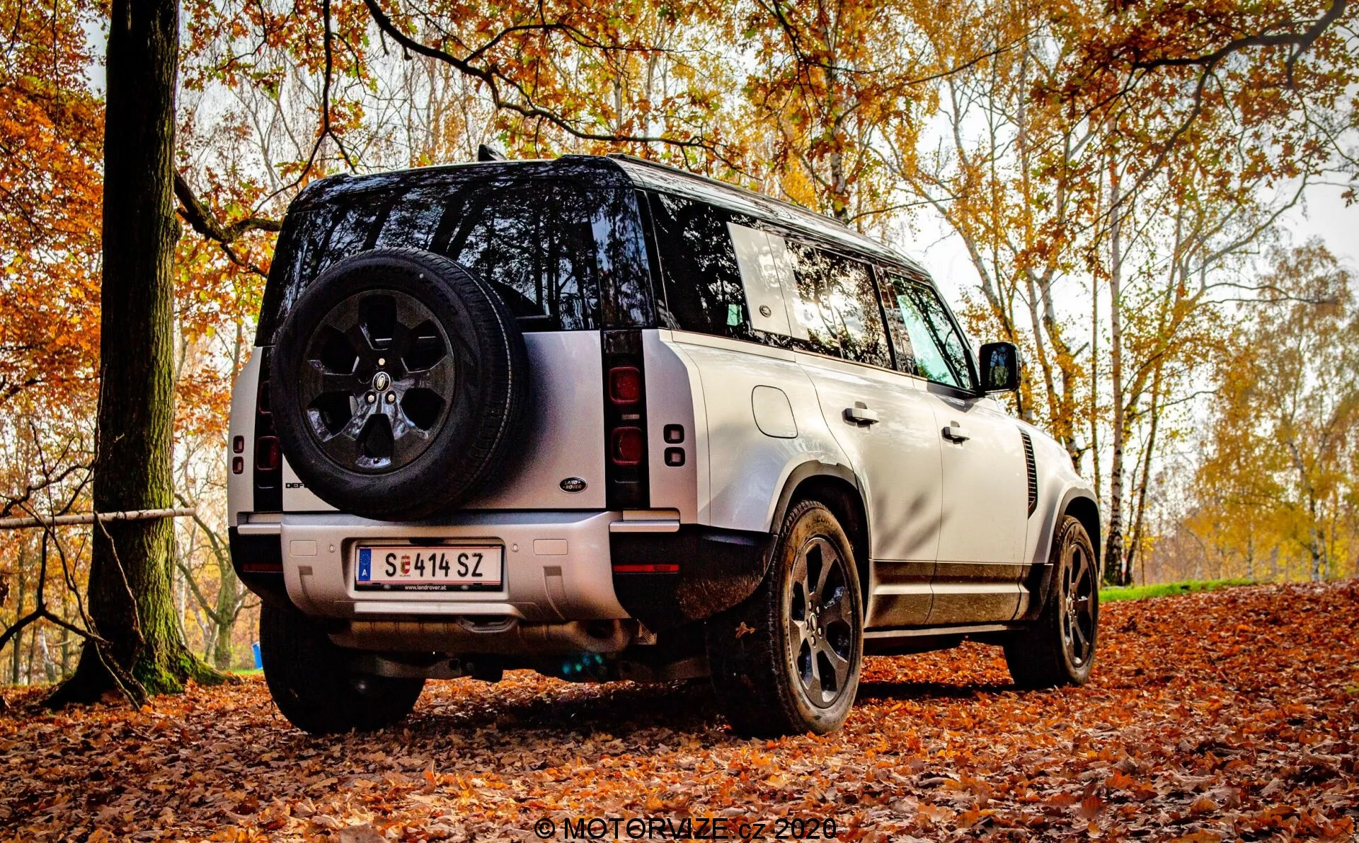 Vista traseira de três quartos de um SUV moderno estacionado em uma trilha de floresta outonal coberta de folhas, destacando a altura e a distância do solo do veículo para capacidades off-road. A imagem revela a traseira, incluindo a porta traseira, parte do teto, a janela traseira e um perfil lateral. As características visíveis incluem o design vertical das luzes traseiras, a roda de liga robusta preta com pneu de espessura, roda sobressalente montada na porta traseira, para-choque traseiro com sensores de estacionamento, pintura em dois tons com teto em contraste, limpador de vidro traseiro, câmera de ré abaixo da placa de licença e degraus laterais para facilitar o acesso à cabine. A carroceria robusta e quadrada transmite luxo com uma linha de caráter forte ao longo das portas laterais. A luz natural e difusa sugere um dia nublado ou filtragem de luz pelo dossel das árvores, melhorando os temas sazonais e ao ar livre.
