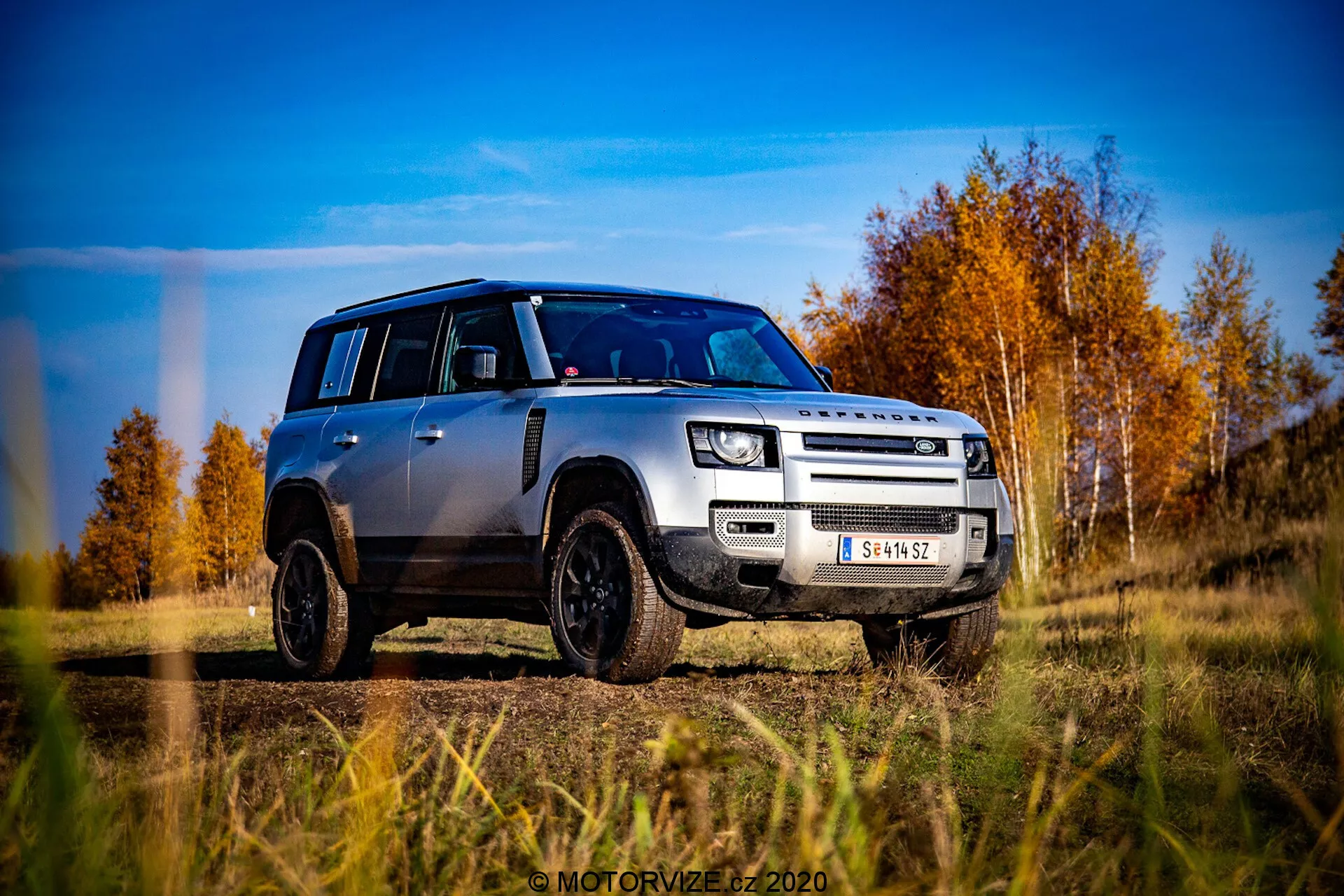 Imagem de um Land Rover Defender 2020 prateado a partir de uma perspectiva ao nível do solo, capturada numa vista frontal oblíqua de três quartos. O veículo destaca a sua grade, capô, para-choques frontal e design das rodas, ligeiramente inclinado para o lado do motorista. O Defender está posicionado em um terreno ao ar livre com fundo de árvores outonais com folhas vermelhas e laranjas sob um céu azul parcialmente nublado. As características notáveis de design incluem sua distinta forma quadrada, faróis de LED modernos, luzes de nevoeiro, grade de duas barras com marca LAND ROVER, placa de deslizamento frontal, jantes de liga preta com pneus off-road e teto preto contrastante com tampas de espelho pretas correspondentes. A foto é altamente nítida e dinâmica, enfatizando a robustez e a estética moderna do veículo.