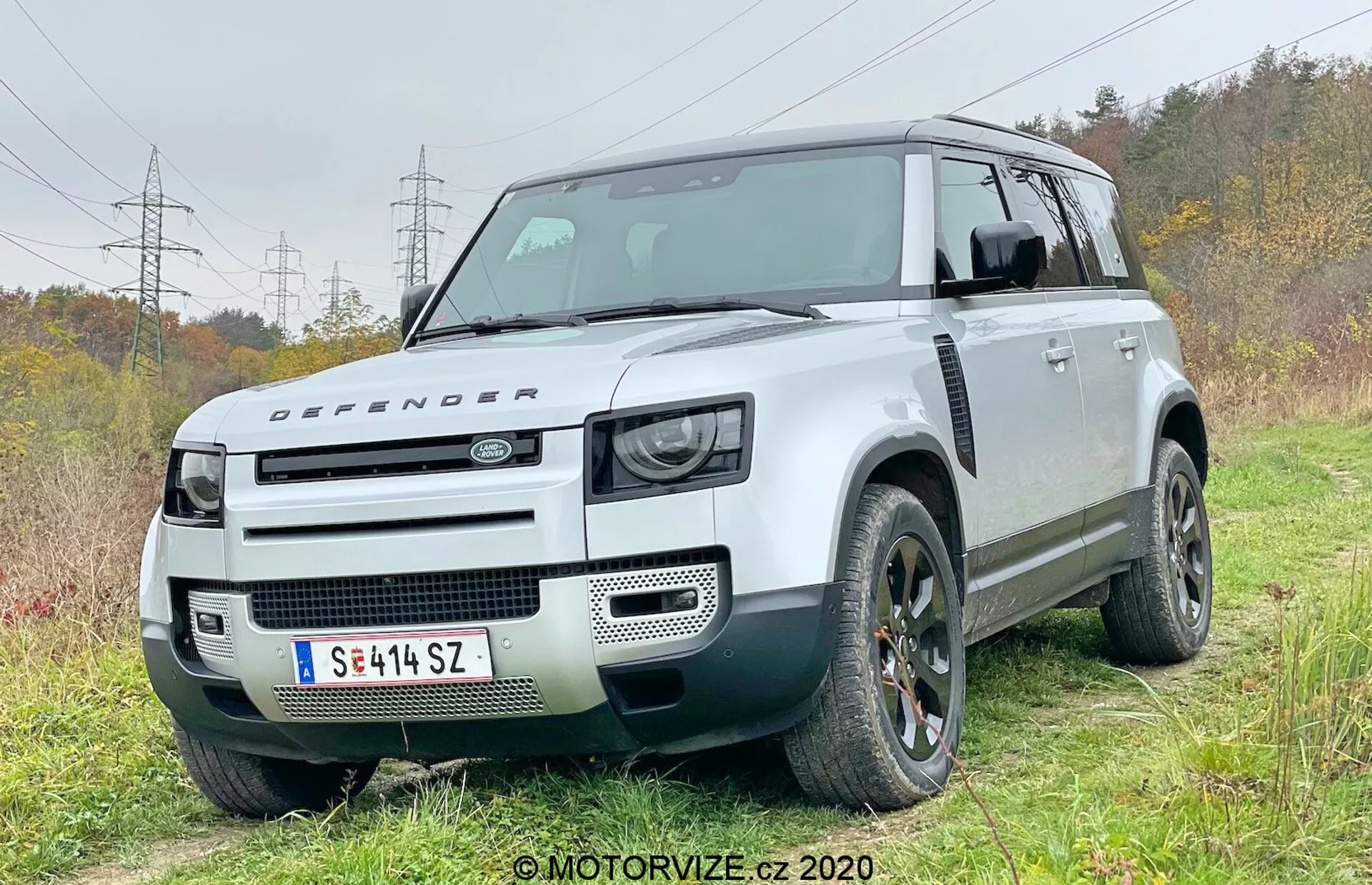 Esta fotografia capta um SUV moderno Land Rover Defender 2020 de um ângulo de frente tridimensional, mostrando o lado do motorista. O veículo está ligeiramente fora de estrada em uma área gramada com uma pista de terra, enfatizando suas capacidades off-road. O design distintivo e quadrado apresenta uma alta distância ao solo, um esquema de cores em dois tons com carroceria branca e teto preto contrastante. A frente exibe conjuntos de faróis LED inspirados no retro, um capô com o emblema Defender e uma grade frontal minimalista, destacando sua reputação robusta todo-o-terreno. As características técnicas visíveis incluem sensores de estacionamento dianteiros integrados no para-choque, sugerindo sistemas avançados de assistência ao motorista. Os robustos arcos das rodas acomodam grandes pneus off-road, e elementos de design como rodas de liga e design exclusivo da grade são destacados.