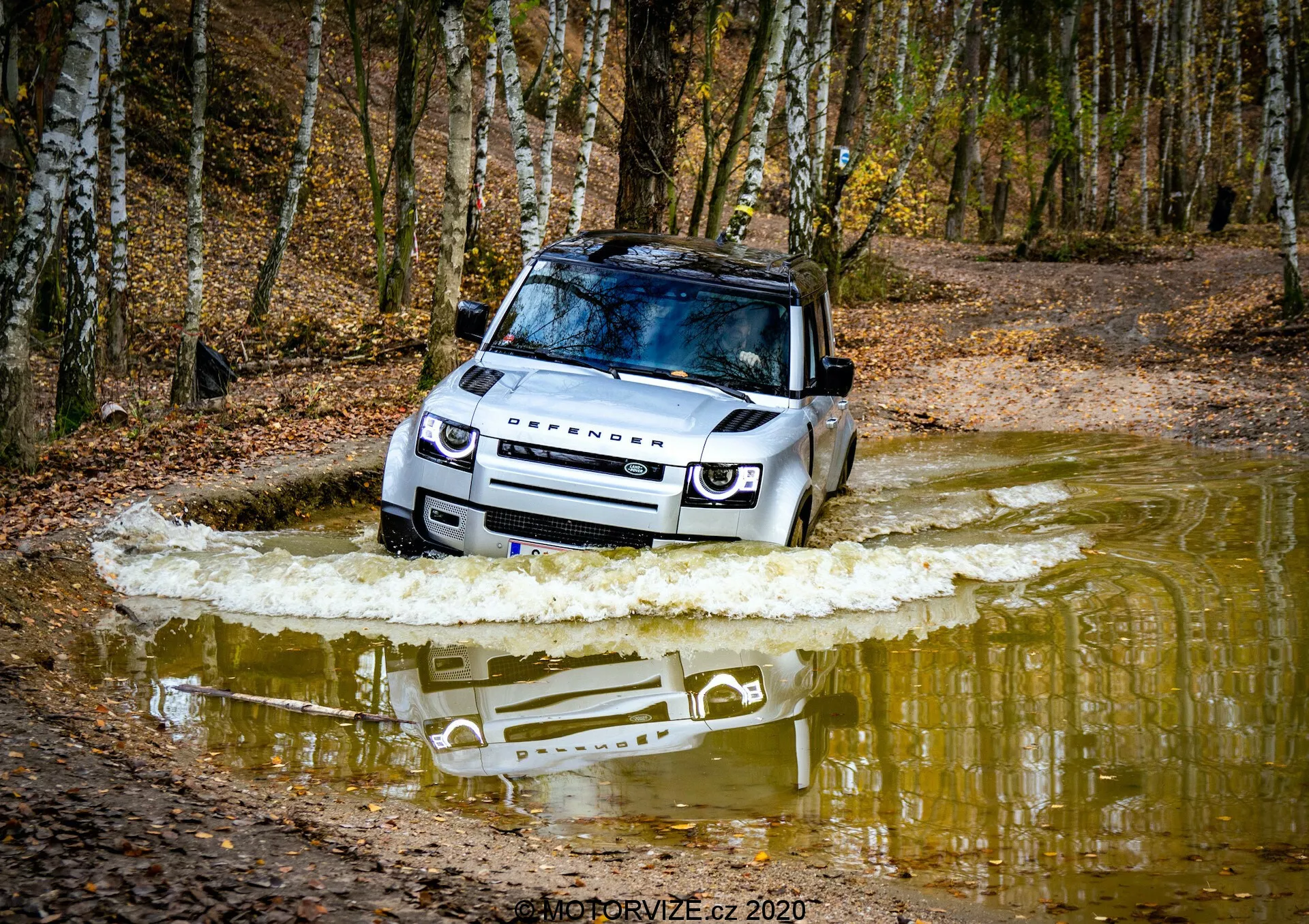 Foto em ângulo baixo de um Land Rover Defender 2020 atravessando um corpo de água, destacando o lado frontal direito com grade visível, faróis LED, capô, para-brisa, teto, roda dianteira e porta do motorista. A cena dinâmica captura o deslocamento da água, enfatizando suas capacidades off-road, alta capacidade de travessia, placa de proteção e teto solar panorâmico de luxo. A marca Land Rover é visível no capô, exibindo seu design robusto e versátil.